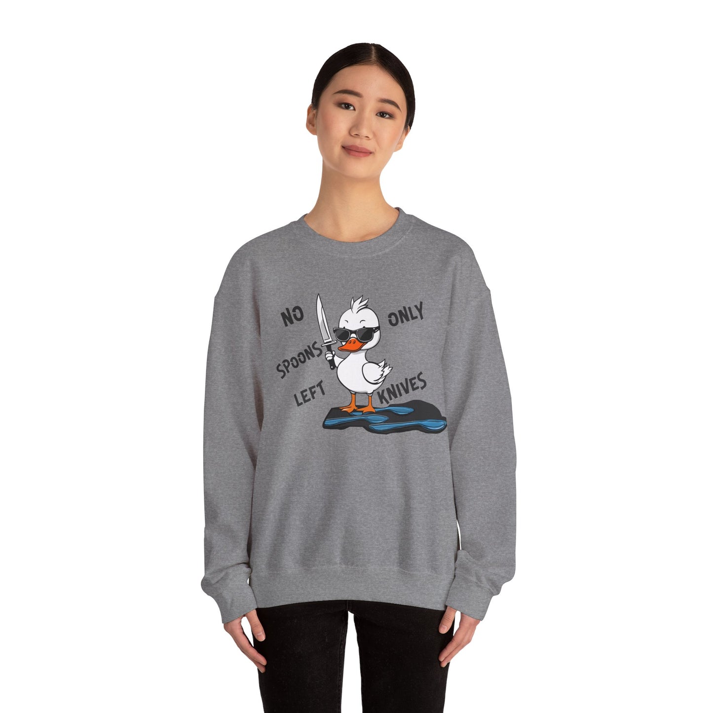 This image shows a woman wearing a graphite heather-colored crewneck sweatshirt with a cartoon design of a duck holding a knife, standing in a puddle of spoons. The text around the duck reads “NO SPOONS LEFT, ONLY KNIVES.” The woman is standing against a plain white background, allowing the sweatshirt’s design to be clearly visible. She is wearing black pants and has a slight smile on her face.