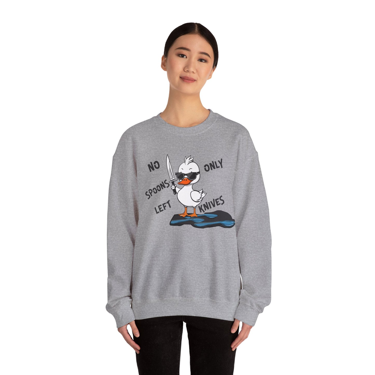 This image shows a woman wearing a sport gray crewneck sweatshirt with a cartoon design of a duck holding a knife, standing in a puddle of spoons. The text around the duck reads “NO SPOONS LEFT, ONLY KNIVES.” The woman is standing against a plain white background, allowing the sweatshirt’s design to be clearly visible. She is wearing black pants and has a slight smile on her face.