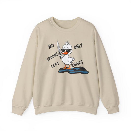 This image shows a sand colored crewneck sweatshirt with a cartoon design of a duck holding a knife, standing in a puddle of spoons. The text around the duck reads “NO SPOONS LEFT, ONLY KNIVES.” The sweatshirt is displayed flat against a plain white background, clearly showcasing the humorous design and text.