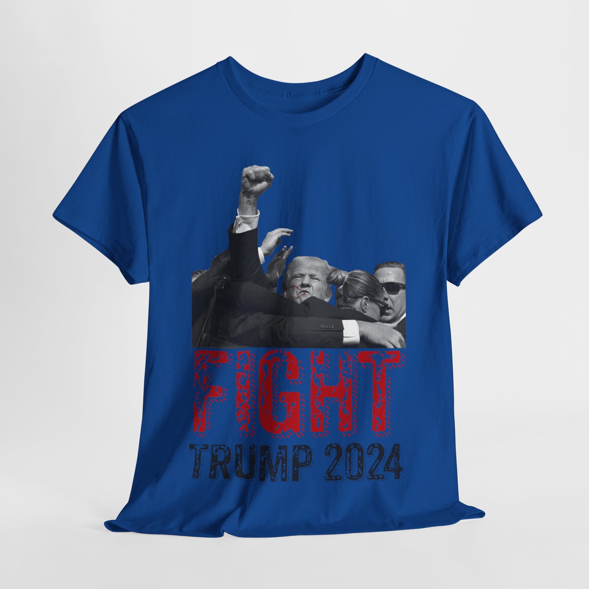 This image displays a royal blue colored, unisex, heavy cotton t-shirt with a graphic design on the front. The design features a black-and-white photograph of former President Donald Trump raising his fist, being shielded by brave men and women. Below the photograph, the words “FIGHT” and “TRUMP 2024” are printed in bold letters, with “FIGHT” in a distressed red font and “TRUMP 2024” in black. The t-shirt is laid out on a plain white background, showcasing the design prominently.