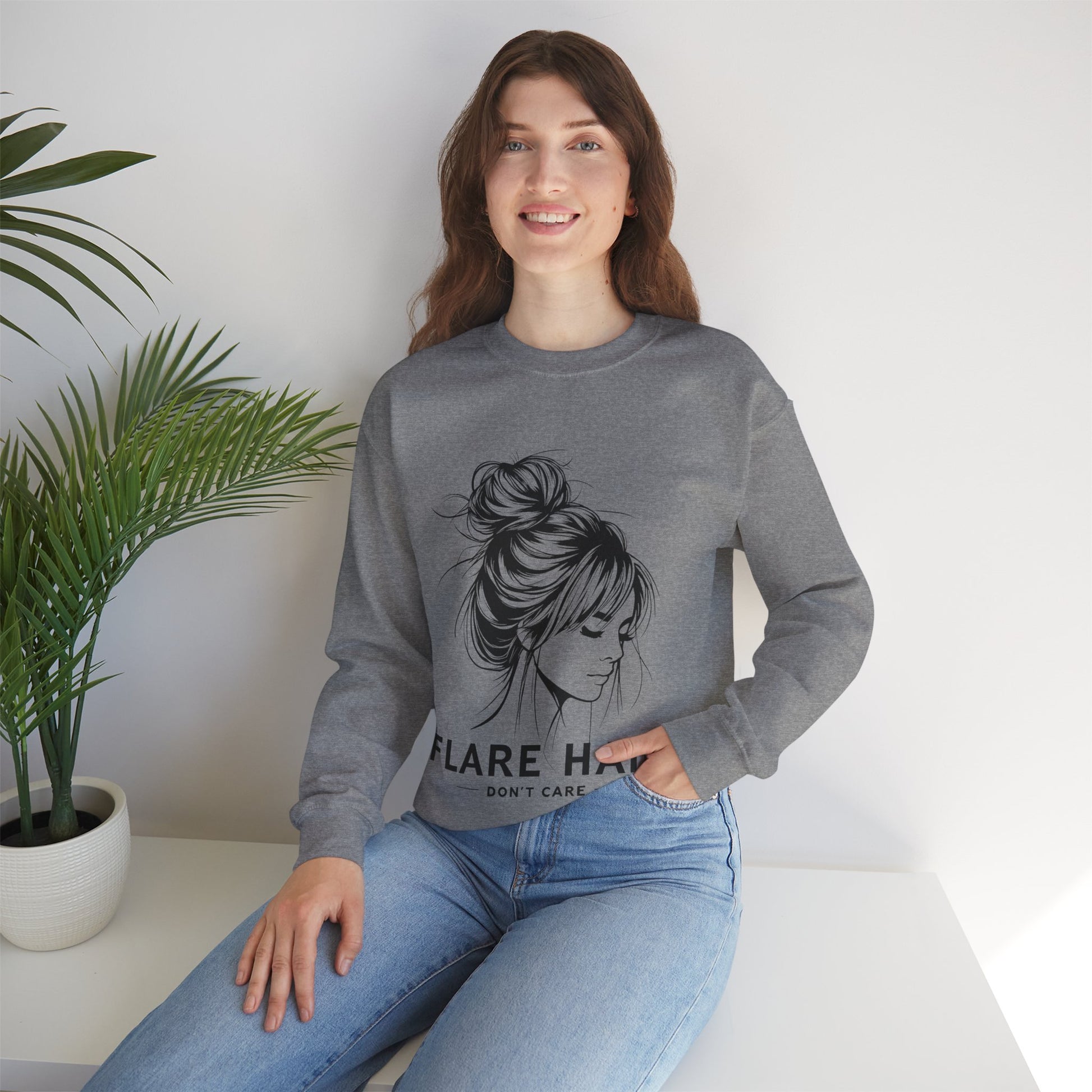 This image features a woman wearing a heather gray crewneck sweatshirt with the text “FLARE HAIR DON’T CARE” printed on the front. The design includes an illustration of a woman with her hair in a messy bun. She is sitting on a white surface with her hand resting on her knee, and there is a potted plant to her left. The background is plain, highlighting the sweatshirt’s design and the casual outfit.