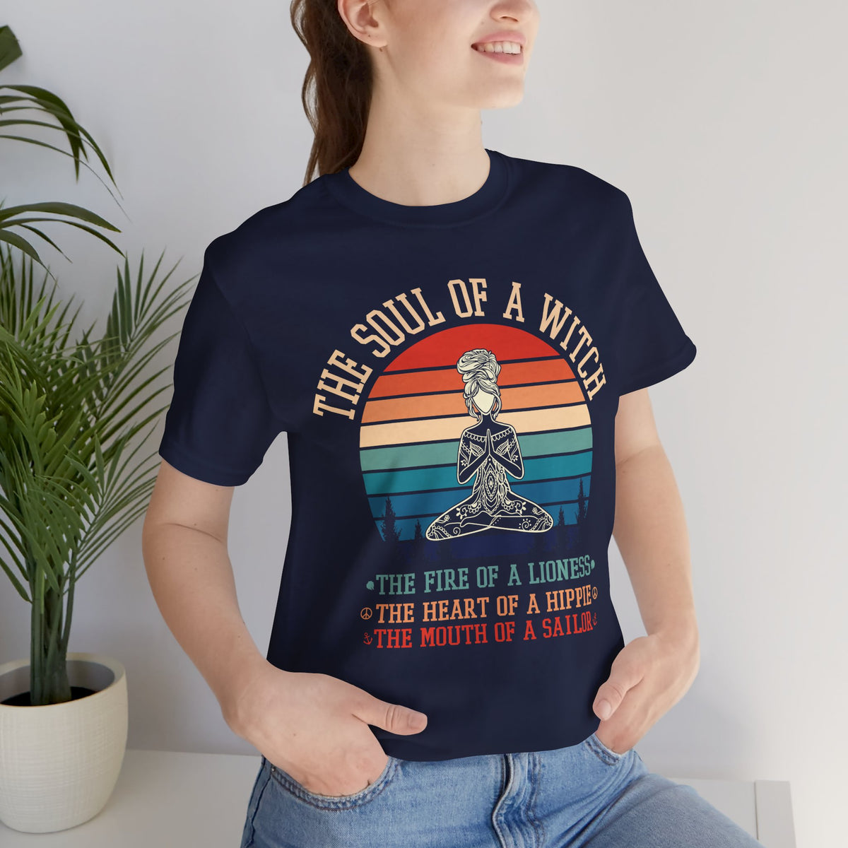 A woman modeling a navy short-sleeve t-shirt with a retro sunset graphic of a woman in lotus pose drawn in mandala linework, reading ‘THE SOUL OF A WITCH, THE FIRE OF A LIONESS, THE HEART OF A HIPPIE, THE MOUTH OF A SAILOR’.