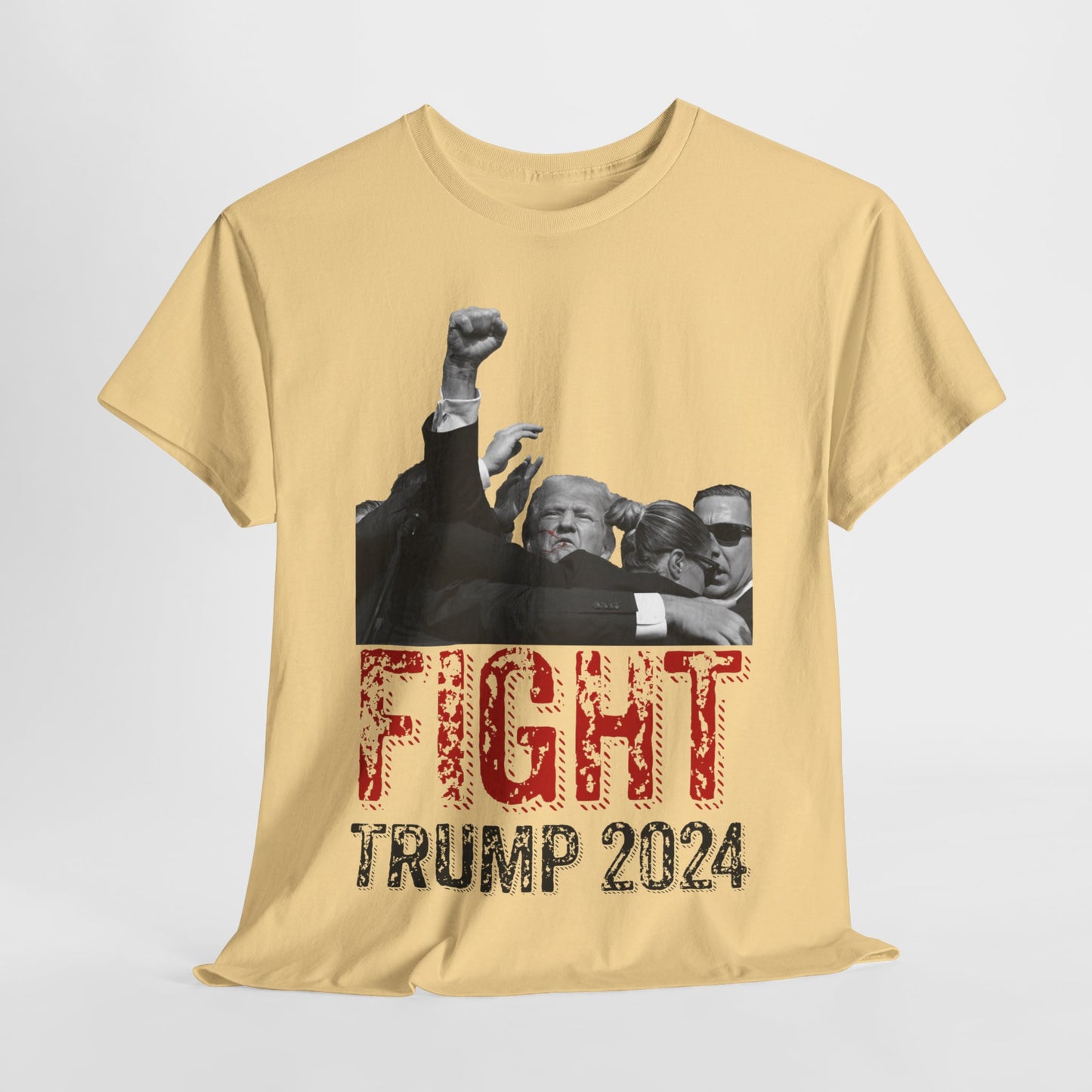 This image displays a yellow haze colored, unisex, heavy cotton t-shirt with a graphic design on the front. The design features a black-and-white photograph of former President Donald Trump raising his fist, being shielded by brave men and women. Below the photograph, the words “FIGHT” and “TRUMP 2024” are printed in bold letters, with “FIGHT” in a distressed red font and “TRUMP 2024” in black. The t-shirt is laid out on a plain white background, showcasing the design prominently.