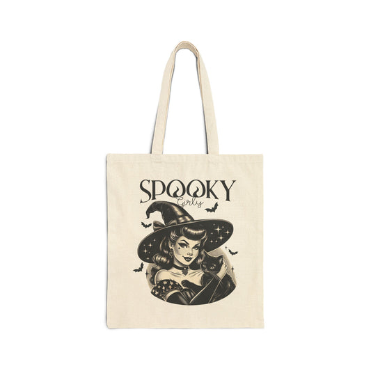 A beige tote bag featuring a vintage-style illustration of a witch with a cat. The witch has wavy hair, wears a wide-brimmed hat, and is surrounded by bats. The text above reads ‘Spooky Girly’ in a playful font