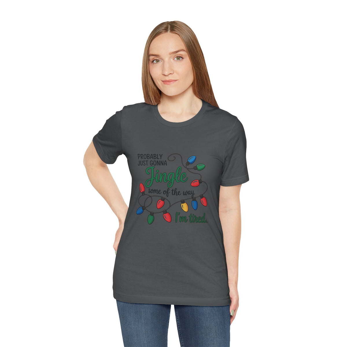 A woman modeling an asphalt short-sleeve t-shirt with a Christmas graphic that reads ‘PROBABLY JUST GONNA Jingle some of the way I'm tired.’ with colorful holiday lights, paired with blue jeans against a plain white background.
