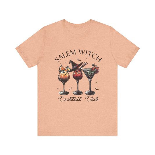 A heather peach colored t-shirt featuring a design with three cocktail glasses, each with a unique drink and garnished with Halloween-themed decorations. The text above the drinks reads “Salem Witch” with a witch’s hat resting on one of the glasses, and the text below reads “Cocktail Club.” The overall vibe is spooky yet fun, perfect for a Halloween-themed gathering or anyone who loves a witchy aesthetic.