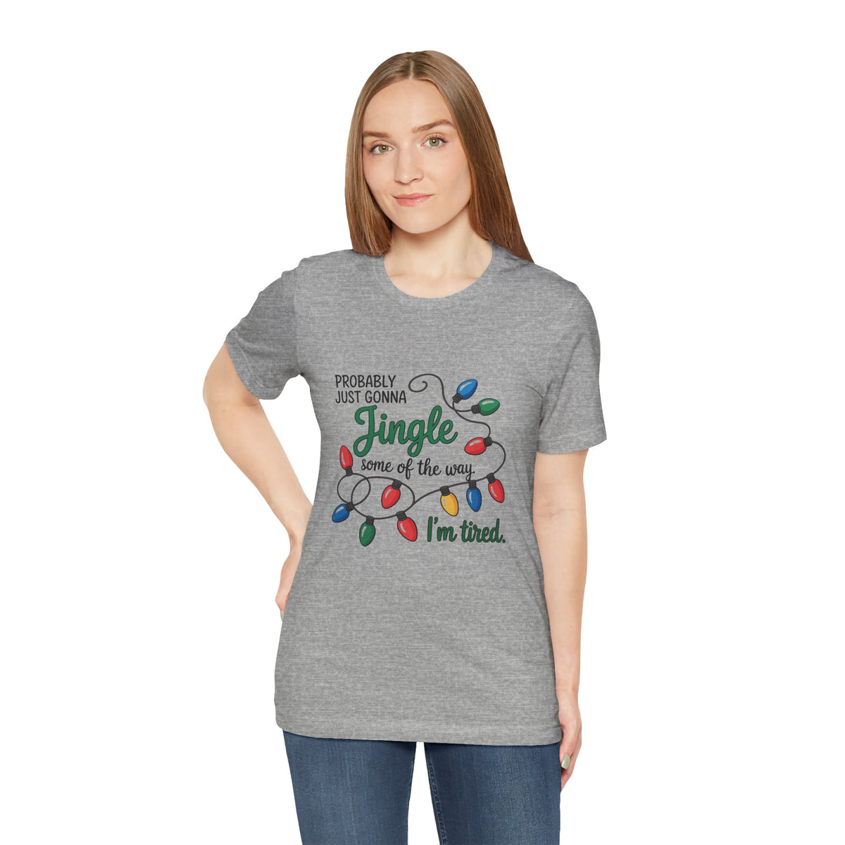 A woman modeling an athletic heather short-sleeve t-shirt with a Christmas graphic that reads ‘PROBABLY JUST GONNA Jingle some of the way I'm tired.’ with colorful holiday lights, paired with blue jeans against a plain white background.