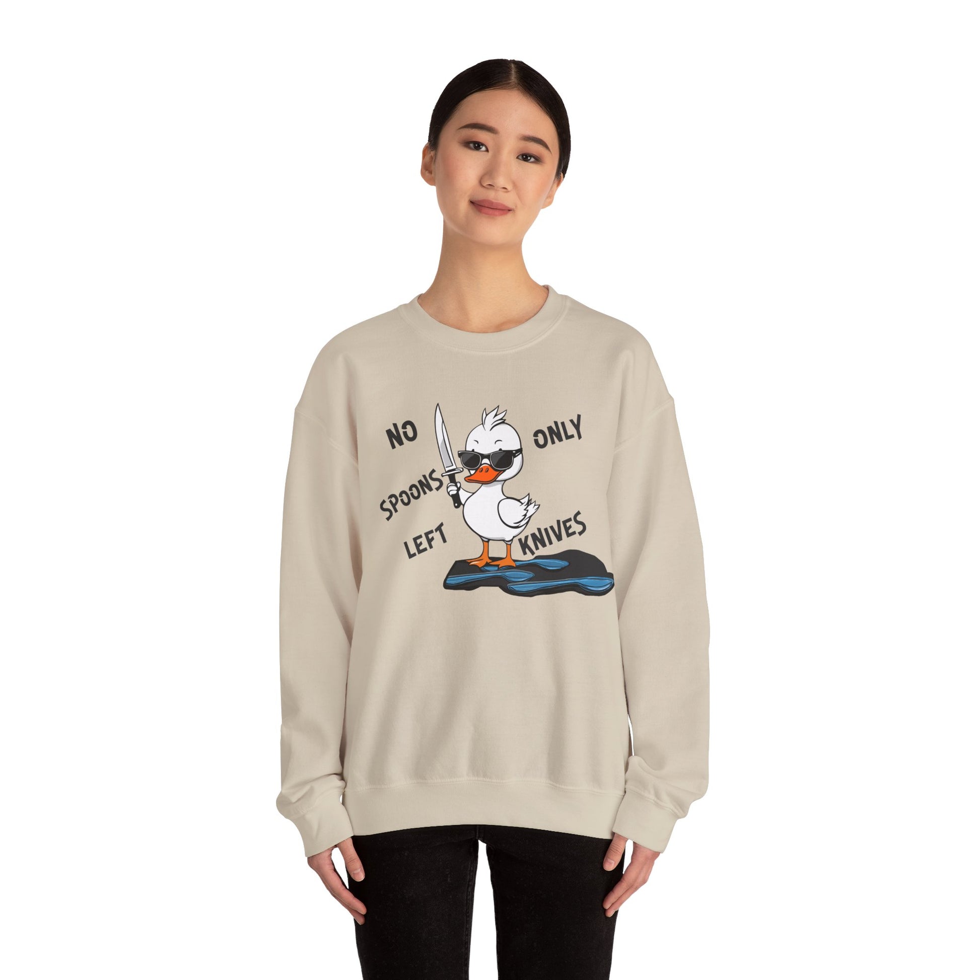 This image shows a woman wearing a sand-colored crewneck sweatshirt with a cartoon design of a duck holding a knife, standing in a puddle of spoons. The text around the duck reads “NO SPOONS LEFT, ONLY KNIVES.” The woman is standing against a plain white background, allowing the sweatshirt’s design to be clearly visible. She is wearing black pants and has a slight smile on her face.