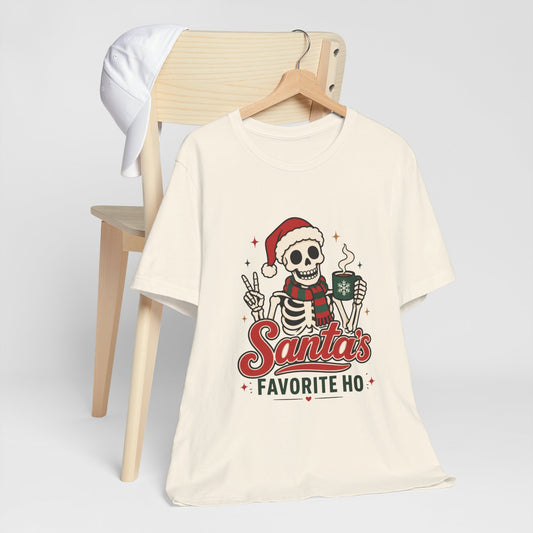 Natural tee printed with a skeleton in a Santa hat and red-and-green striped scarf, giving a peace sign and holding a steaming mug with a snowflake, reading "Santa's Favorite Ho".
