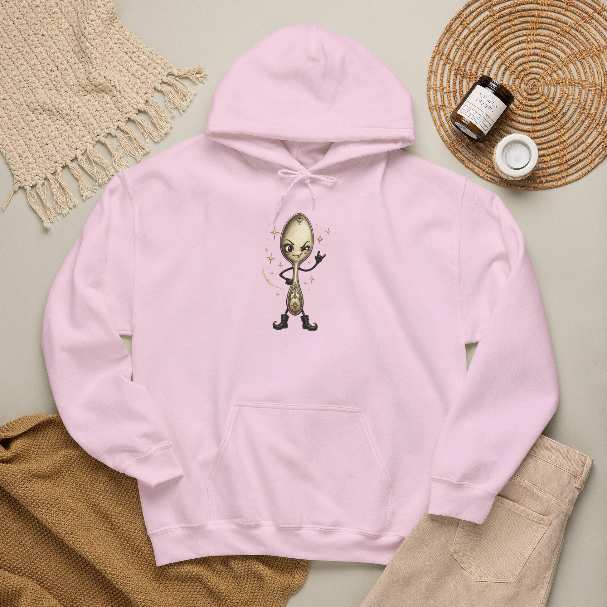A light pink pullover hoodie laid flat with a central graphic of a cartoon spoon in true rock spirit hand. The hoodie is styled in a flat lay with a beige knit blanket, tan pants, a woven mat, and small jars on a neutral background.