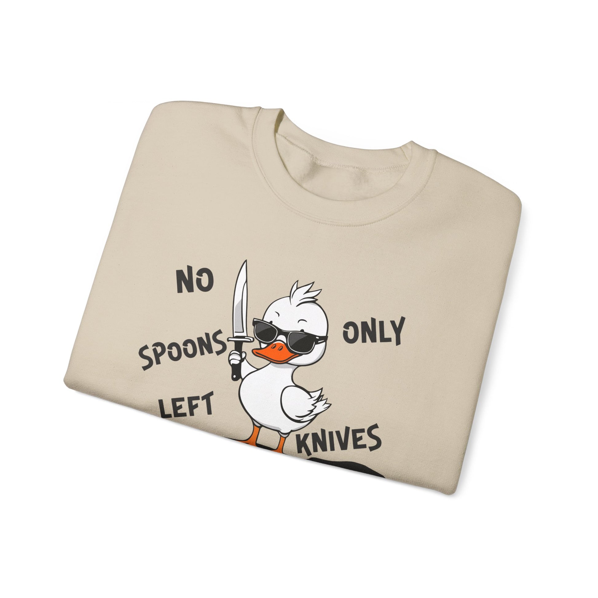 This image shows a sand-colored crewneck sweatshirt folded neatly. The sweatshirt features a cartoon design of a duck holding a knife, standing in a puddle of spoons. The text around the duck reads “NO SPOONS LEFT, ONLY KNIVES.” The sweatshirt is displayed against a plain white background, clearly showcasing the design and text.