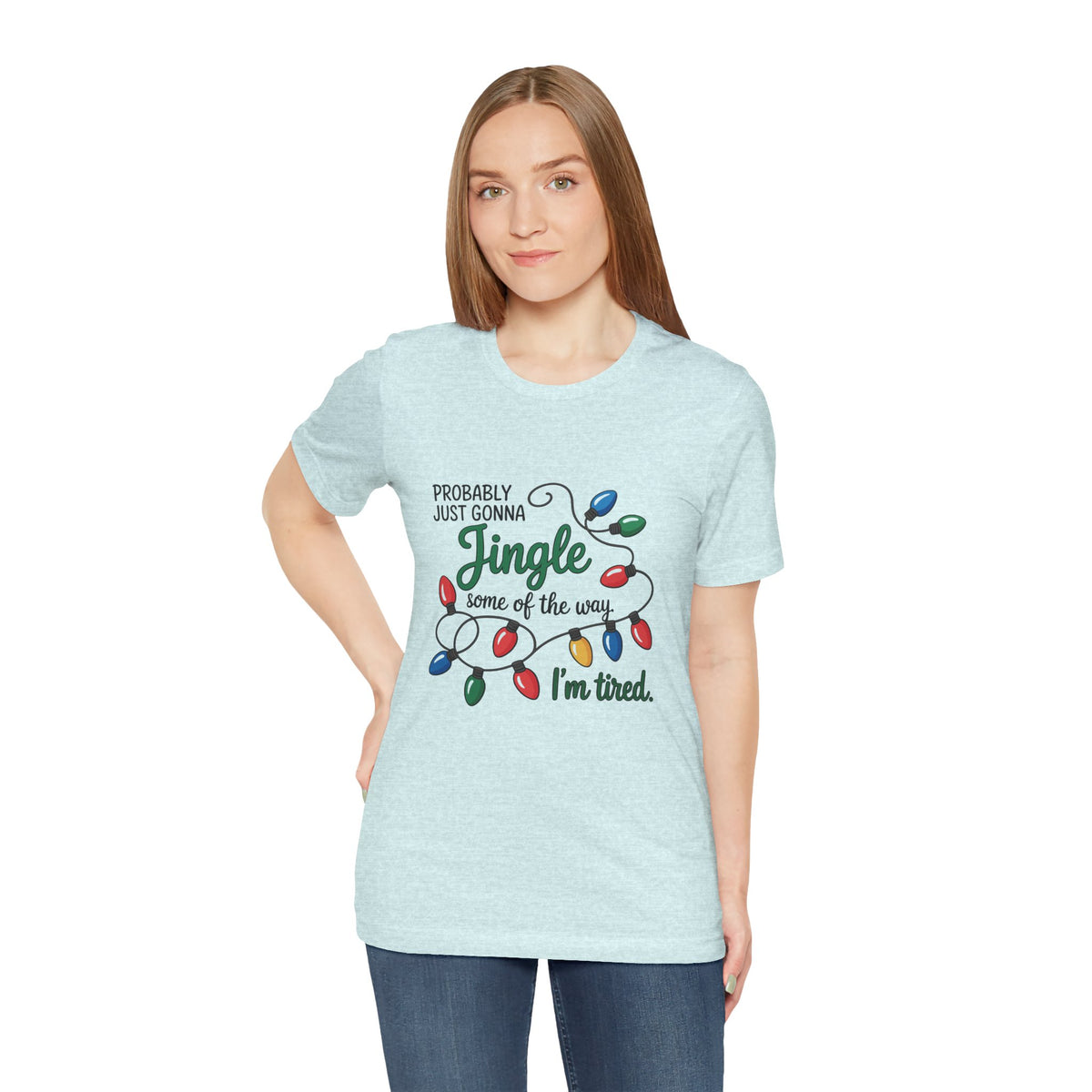 A woman modeling a heather prism ice blue short-sleeve t-shirt with a Christmas graphic that reads ‘PROBABLY JUST GONNA Jingle some of the way I'm tired.’ with colorful holiday lights, paired with blue jeans against a plain white background.