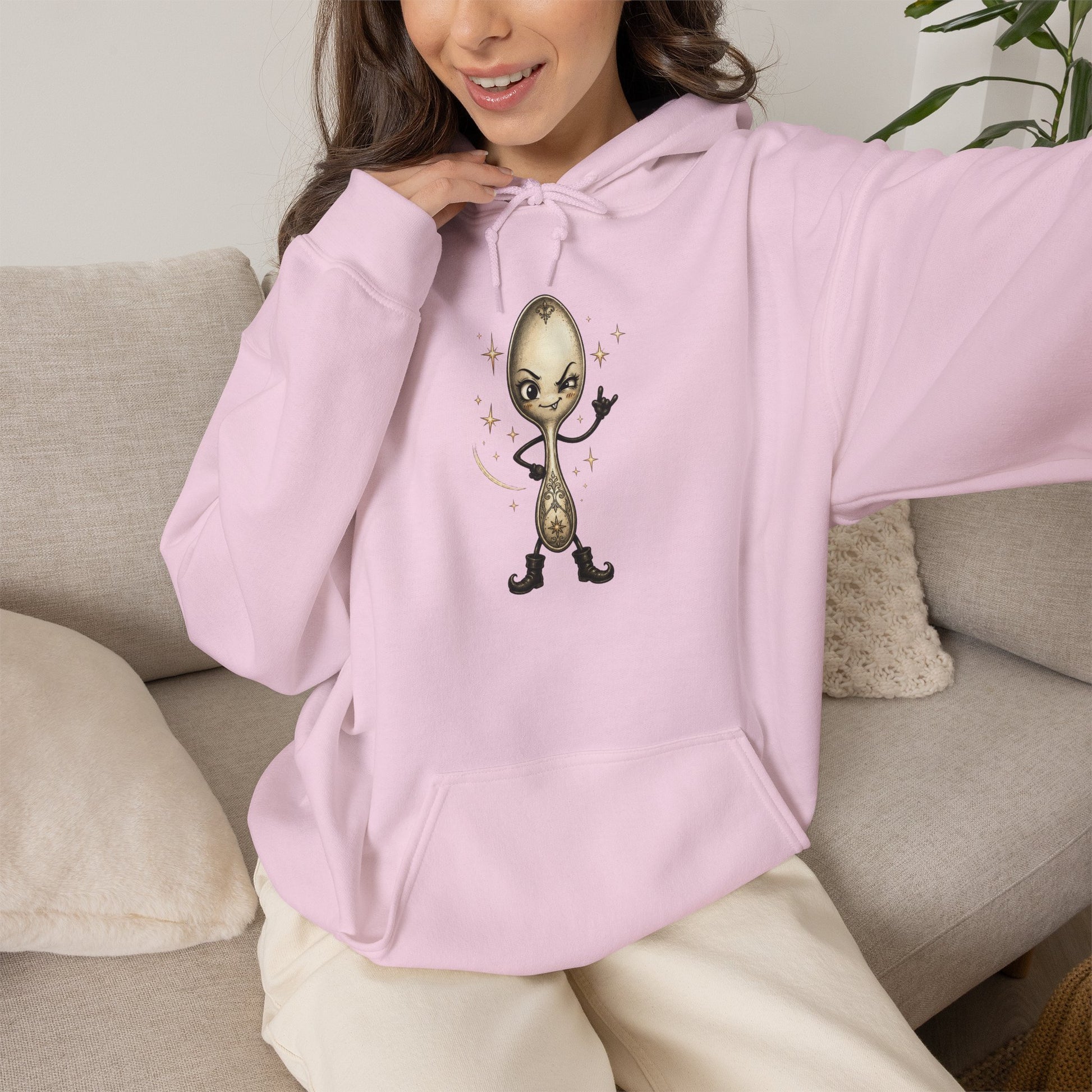 A woman wearing a light pink hoodie with a cartoon spoon graphic on the chest, sitting on a beige couch.