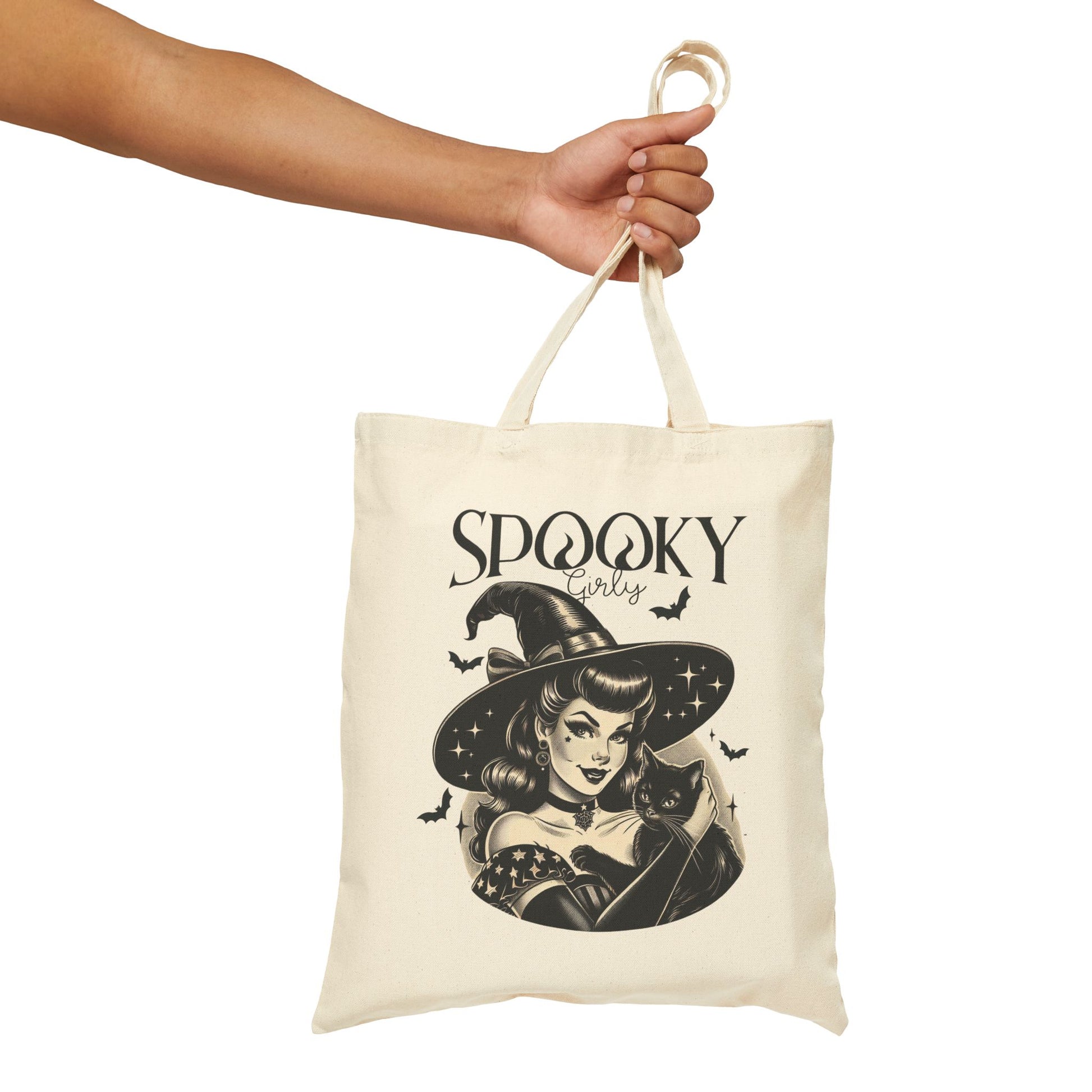 A person holding a beige tote bag with one hand, featuring a vintage-style illustration of a witch and a cat. The design includes the text ‘Spooky Girly’ with bats around the witch’s hat.