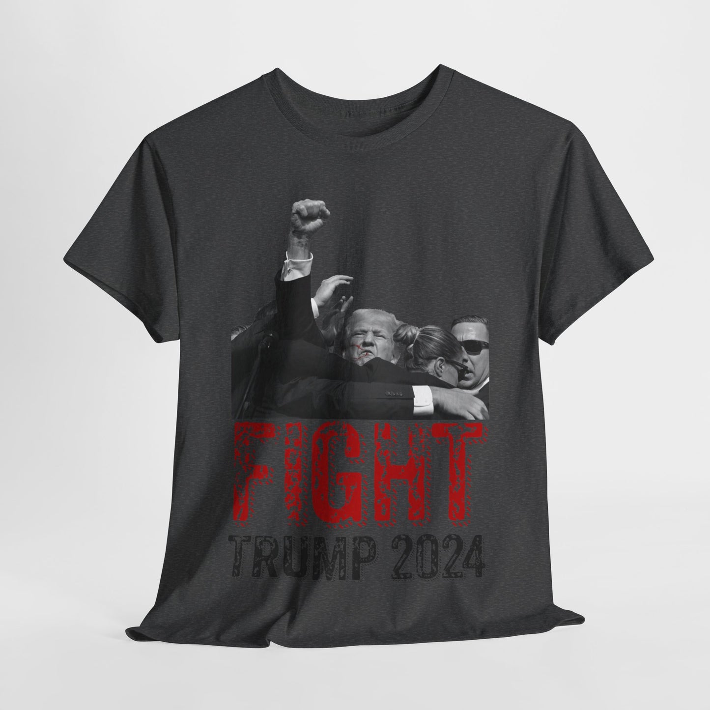 This image displays a dark-heather colored, unisex, heavy cotton t-shirt with a graphic design on the front. The design features a black-and-white photograph of former President Donald Trump raising his fist, being shielded by brave men and women. Below the photograph, the words “FIGHT” and “TRUMP 2024” are printed in bold letters, with “FIGHT” in a distressed red font and “TRUMP 2024” in black. The t-shirt is laid out on a plain white background, showcasing the design prominently.