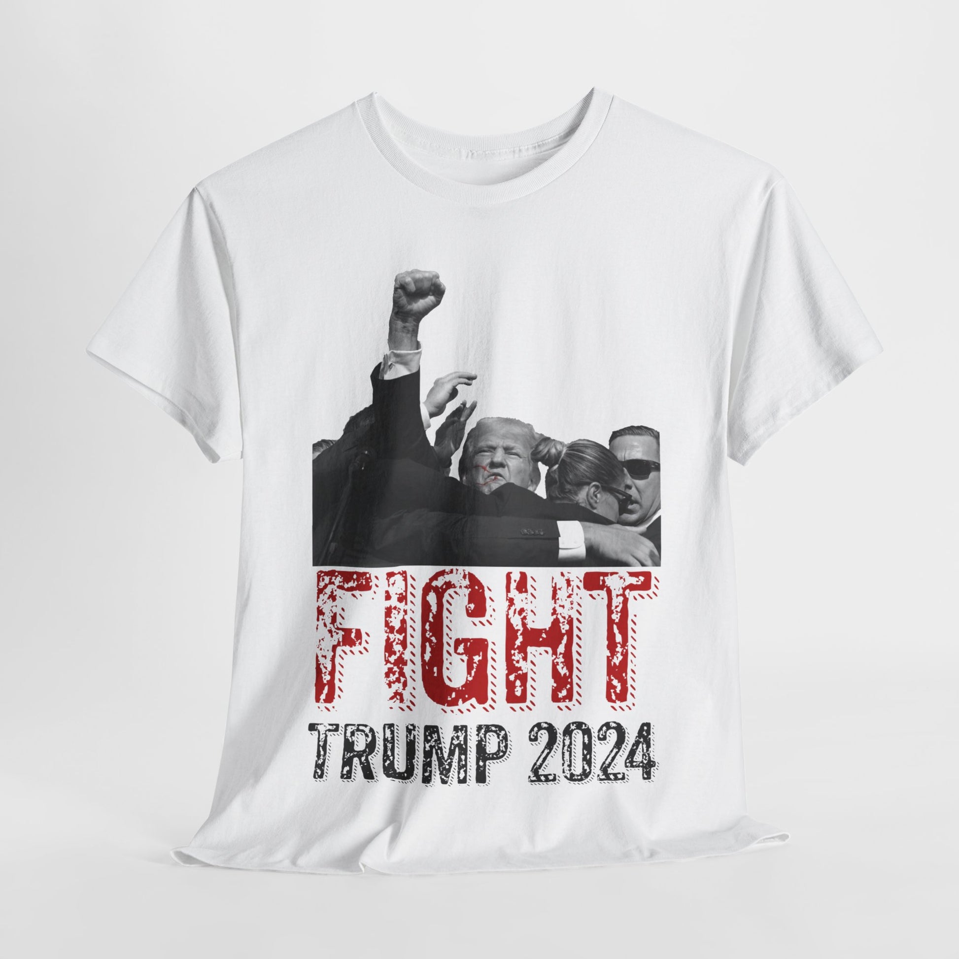 This image displays a white, unisex, heavy cotton t-shirt with a graphic design on the front. The design features a black-and-white photograph of former President Donald Trump raising his fist, being shielded by brave men and women. Below the photograph, the words “FIGHT” and “TRUMP 2024” are printed in bold letters, with “FIGHT” in a distressed red font and “TRUMP 2024” in black. The t-shirt is laid out on a plain white background, showcasing the design prominently.