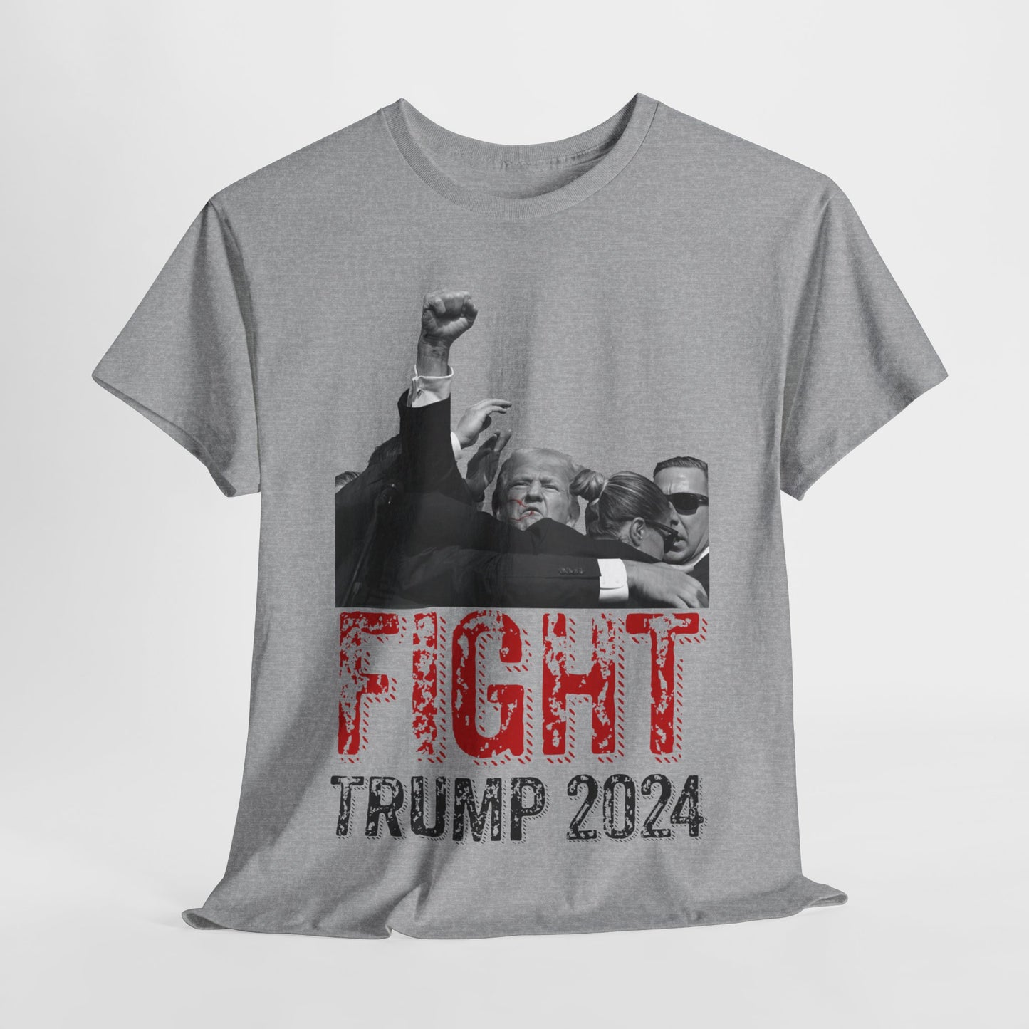 This image displays a sport grey, unisex, heavy cotton t-shirt with a graphic design on the front. The design features a black-and-white photograph of former President Donald Trump raising his fist, being shielded by brave men and women. Below the photograph, the words “FIGHT” and “TRUMP 2024” are printed in bold letters, with “FIGHT” in a distressed red font and “TRUMP 2024” in black. The t-shirt is laid out on a plain white background, showcasing the design prominently.