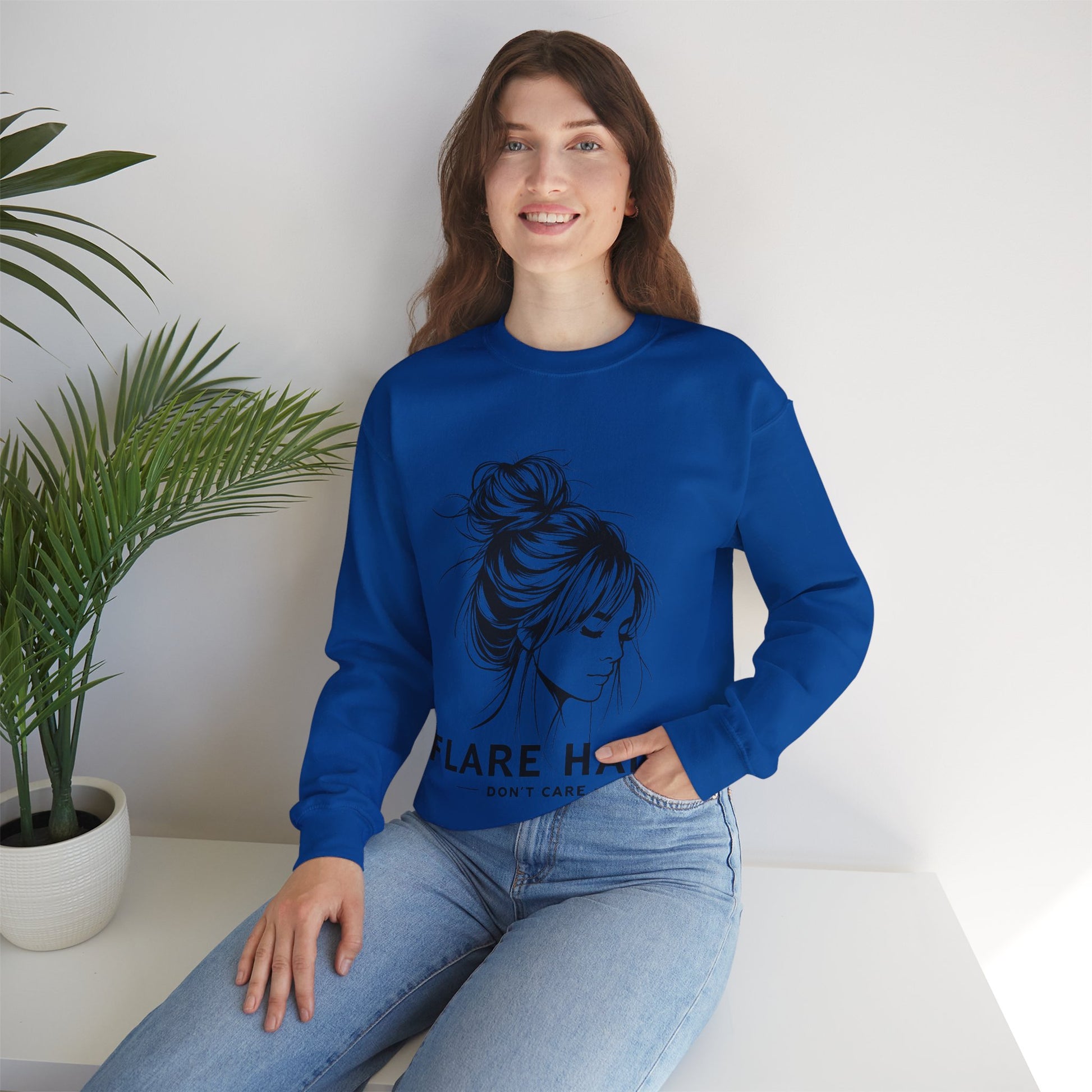 This image features a woman wearing a royal blue crewneck sweatshirt with the text “FLARE HAIR DON’T CARE” printed on the front. The design includes an illustration of a woman with her hair in a messy bun. She is sitting on a white surface with her hand resting on her knee, and there is a potted plant to her left. The background is plain, highlighting the sweatshirt’s design and the casual outfit.