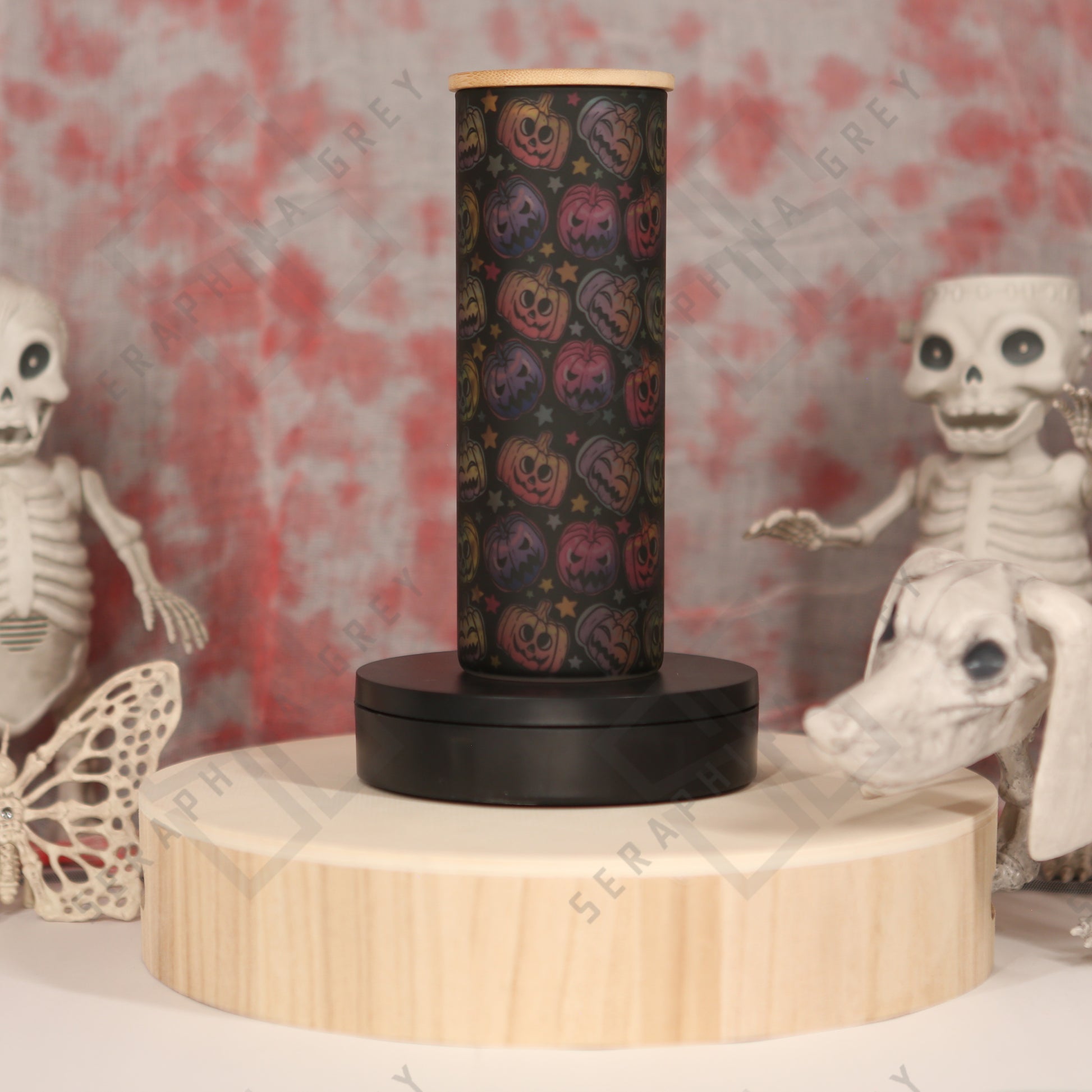 A 25 oz glass tumbler with a colorful pumpkin pattern is displayed on a wooden platform. The tumbler has a black base and a bamboo lid. In the background, there are Halloween-themed decorations, including skeleton figures and a butterfly. The scene has a spooky and festive atmosphere.