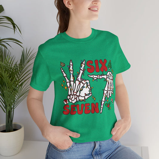 A woman wearing a heather kelly short-sleeve t-shirt with a graphic of skeleton hands wrapped in Christmas lights, with a small tree, ornament, mistletoe and a present, and the words ‘SIX SEVEN’ in red.