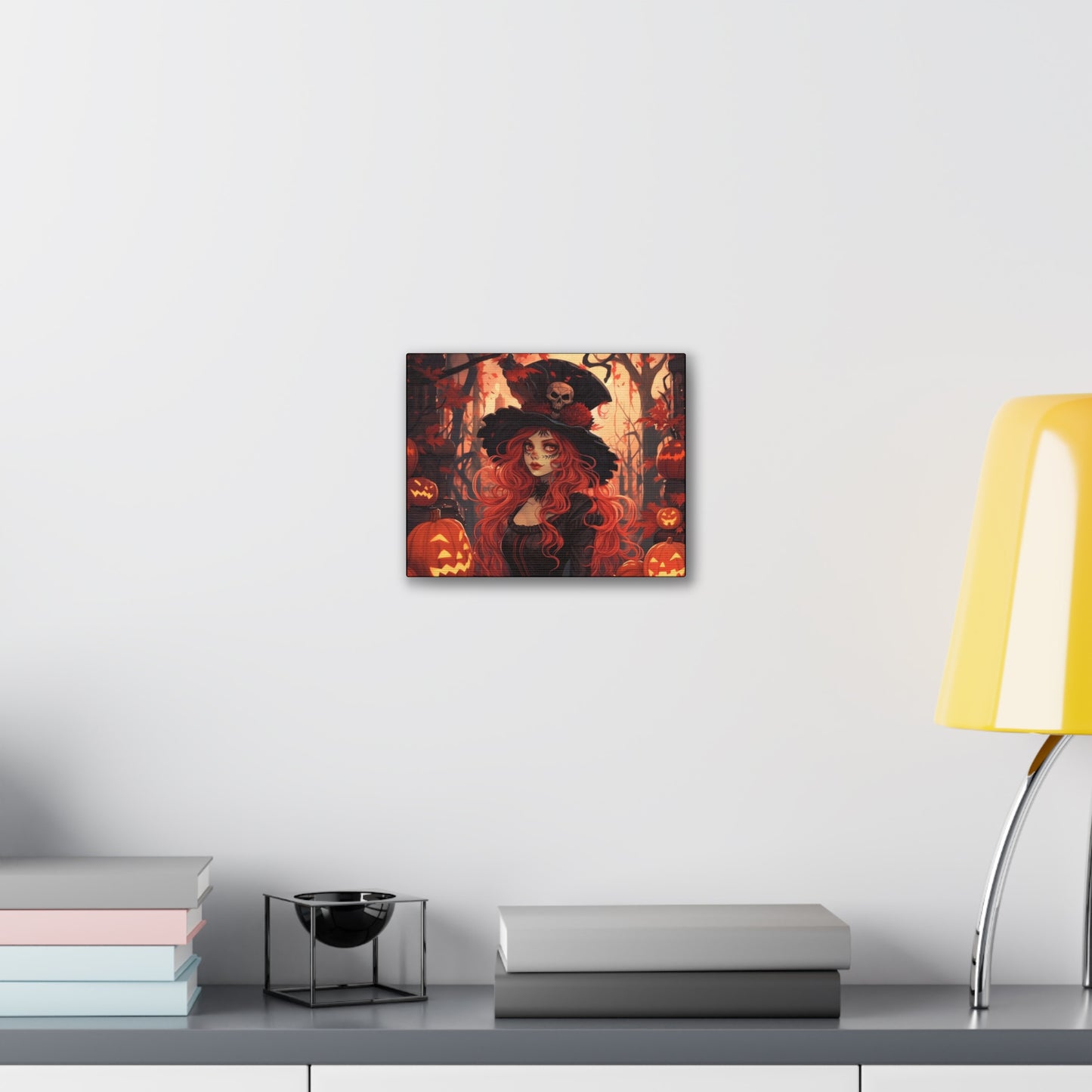 A canvas print titled “Pumpkin Queen” hangs on a white wall above a modern shelf. The shelf holds stacked books, a small black bowl, and is next to a yellow lamp. The artwork depicts a red-haired witch with a skull-adorned black hat, surrounded by carved pumpkins and autumn leaves in a spooky forest, creating a Halloween-themed atmosphere.