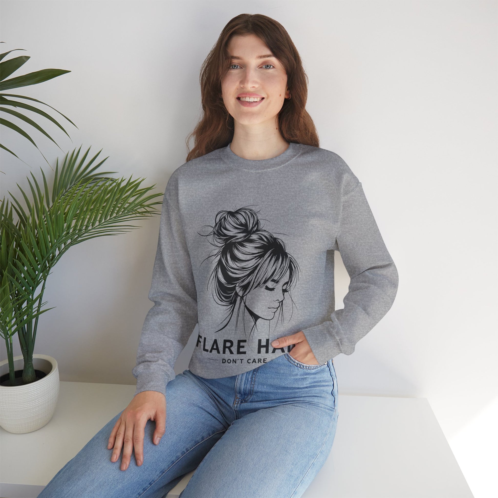 This image features a woman wearing a sport gray crewneck sweatshirt with the text “FLARE HAIR DON’T CARE” printed on the front. The design includes an illustration of a woman with her hair in a messy bun. She is sitting on a white surface with her hand resting on her knee, and there is a potted plant to her left. The background is plain, highlighting the sweatshirt’s design and the casual outfit.