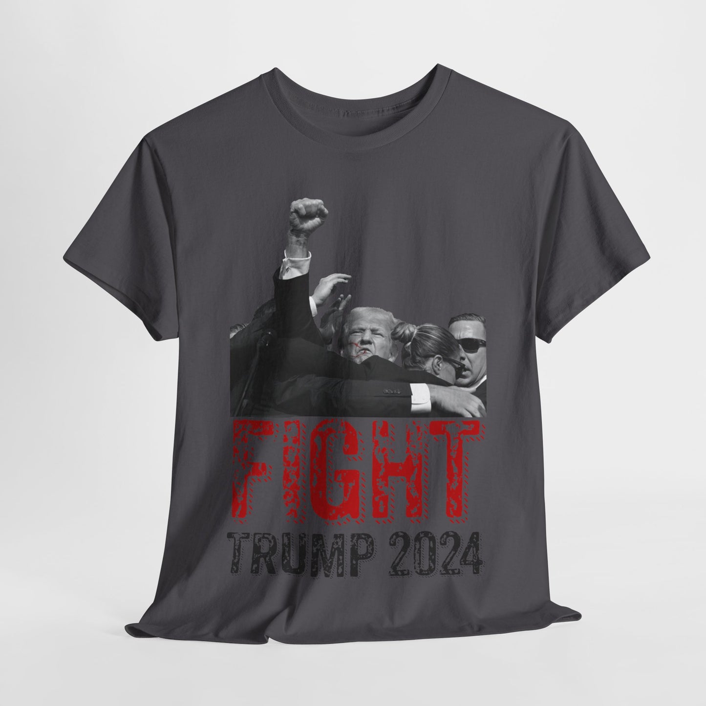 This image displays a charcoal colored, unisex, heavy cotton t-shirt with a graphic design on the front. The design features a black-and-white photograph of former President Donald Trump raising his fist, being shielded by brave men and women. Below the photograph, the words “FIGHT” and “TRUMP 2024” are printed in bold letters, with “FIGHT” in a distressed red font and “TRUMP 2024” in black. The t-shirt is laid out on a plain white background, showcasing the design prominently.