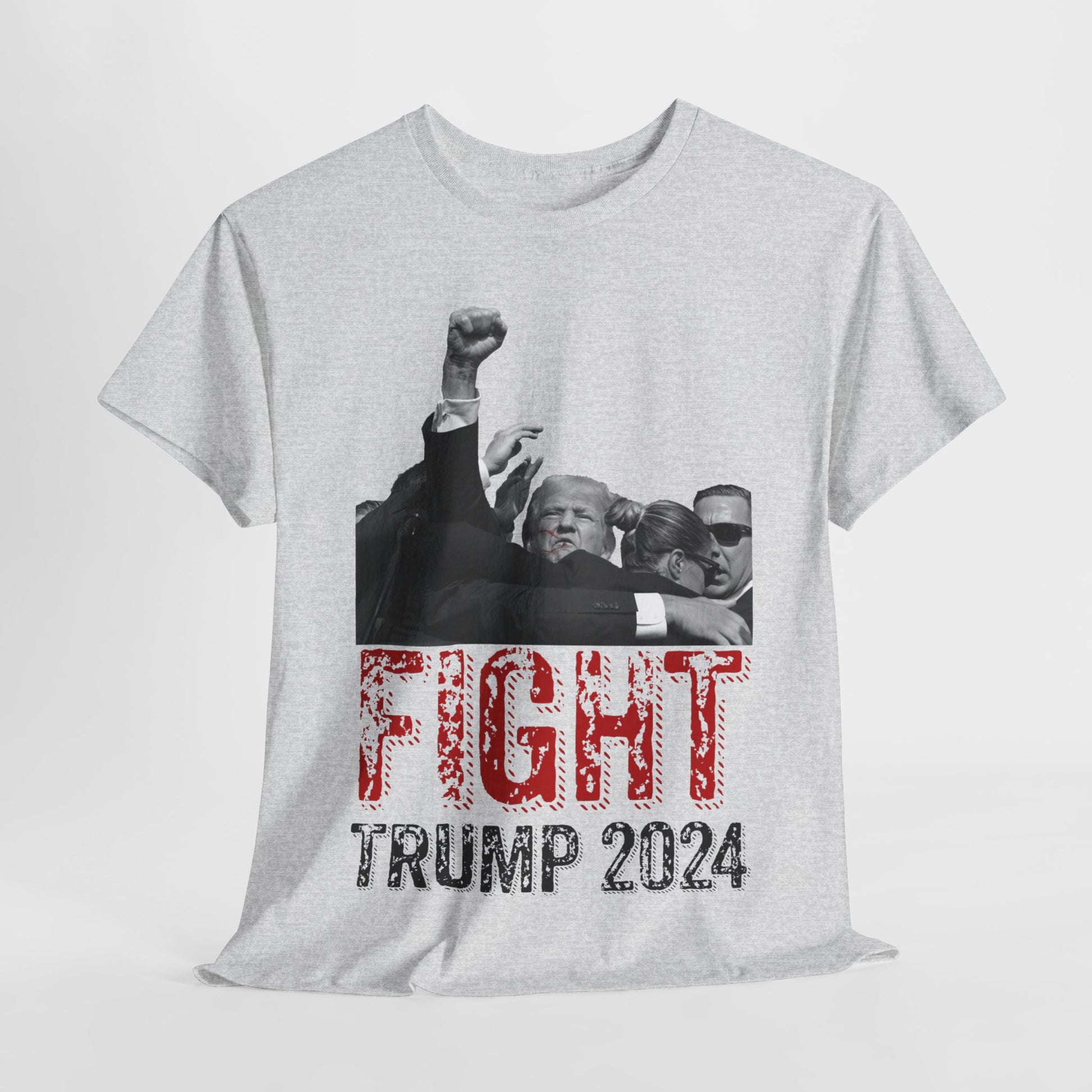 This image displays an ash-colored, unisex, heavy cotton t-shirt with a graphic design on the front. The design features a black-and-white photograph of former President Donald Trump raising his fist, being shielded by brave men and women. Below the photograph, the words “FIGHT” and “TRUMP 2024” are printed in bold letters, with “FIGHT” in a distressed red font and “TRUMP 2024” in black. The t-shirt is laid out on a plain white background, showcasing the design prominently.