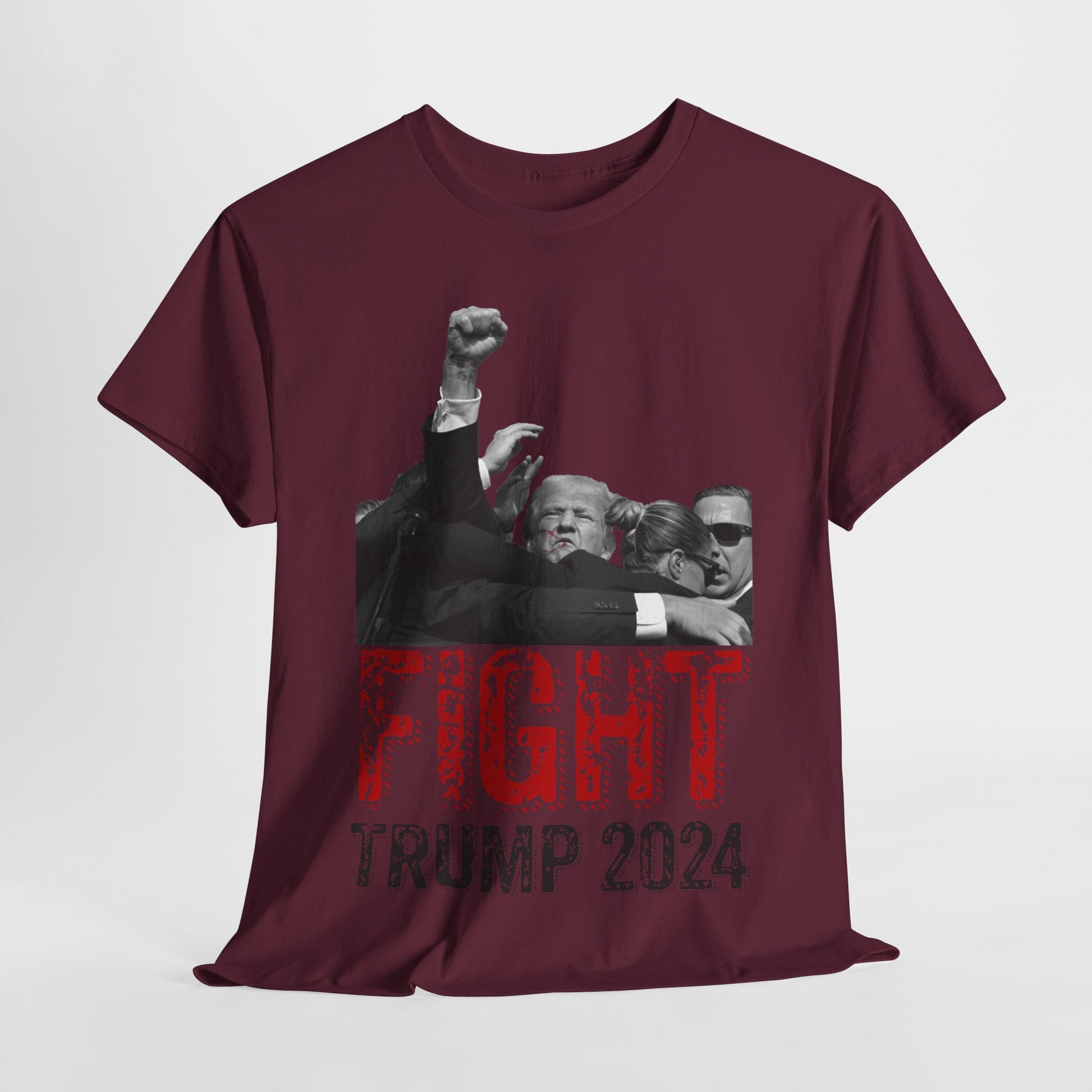 This image displays a maroon colored, unisex, heavy cotton t-shirt with a graphic design on the front. The design features a black-and-white photograph of former President Donald Trump raising his fist, being shielded by brave men and women. Below the photograph, the words “FIGHT” and “TRUMP 2024” are printed in bold letters, with “FIGHT” in a distressed red font and “TRUMP 2024” in black. The t-shirt is laid out on a plain white background, showcasing the design prominently.