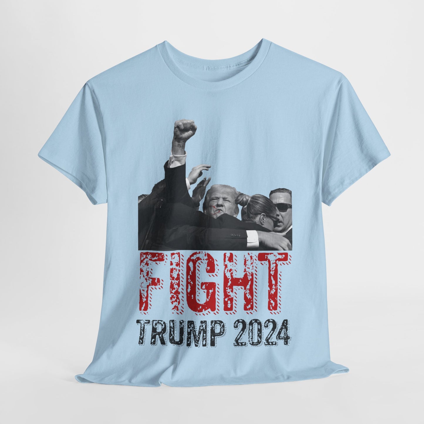 This image displays a light blue colored, unisex, heavy cotton t-shirt with a graphic design on the front. The design features a black-and-white photograph of former President Donald Trump raising his fist, being shielded by brave men and women. Below the photograph, the words “FIGHT” and “TRUMP 2024” are printed in bold letters, with “FIGHT” in a distressed red font and “TRUMP 2024” in black. The t-shirt is laid out on a plain white background, showcasing the design prominently.