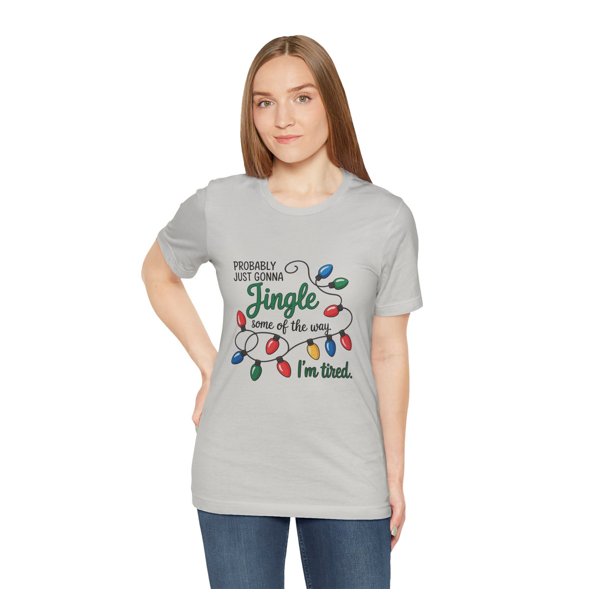 A woman modeling a silver short-sleeve t-shirt with a Christmas graphic that reads ‘PROBABLY JUST GONNA Jingle some of the way I'm tired.’ with colorful holiday lights, paired with blue jeans against a plain white background.