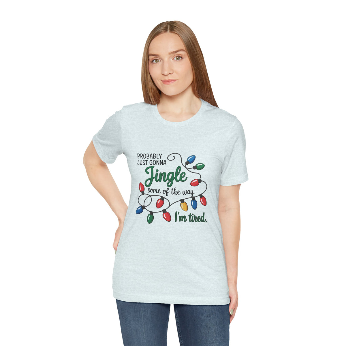 A woman modeling a heather ice blue short-sleeve t-shirt with a Christmas graphic that reads ‘PROBABLY JUST GONNA Jingle some of the way I'm tired.’ with colorful holiday lights, paired with blue jeans against a plain white background.