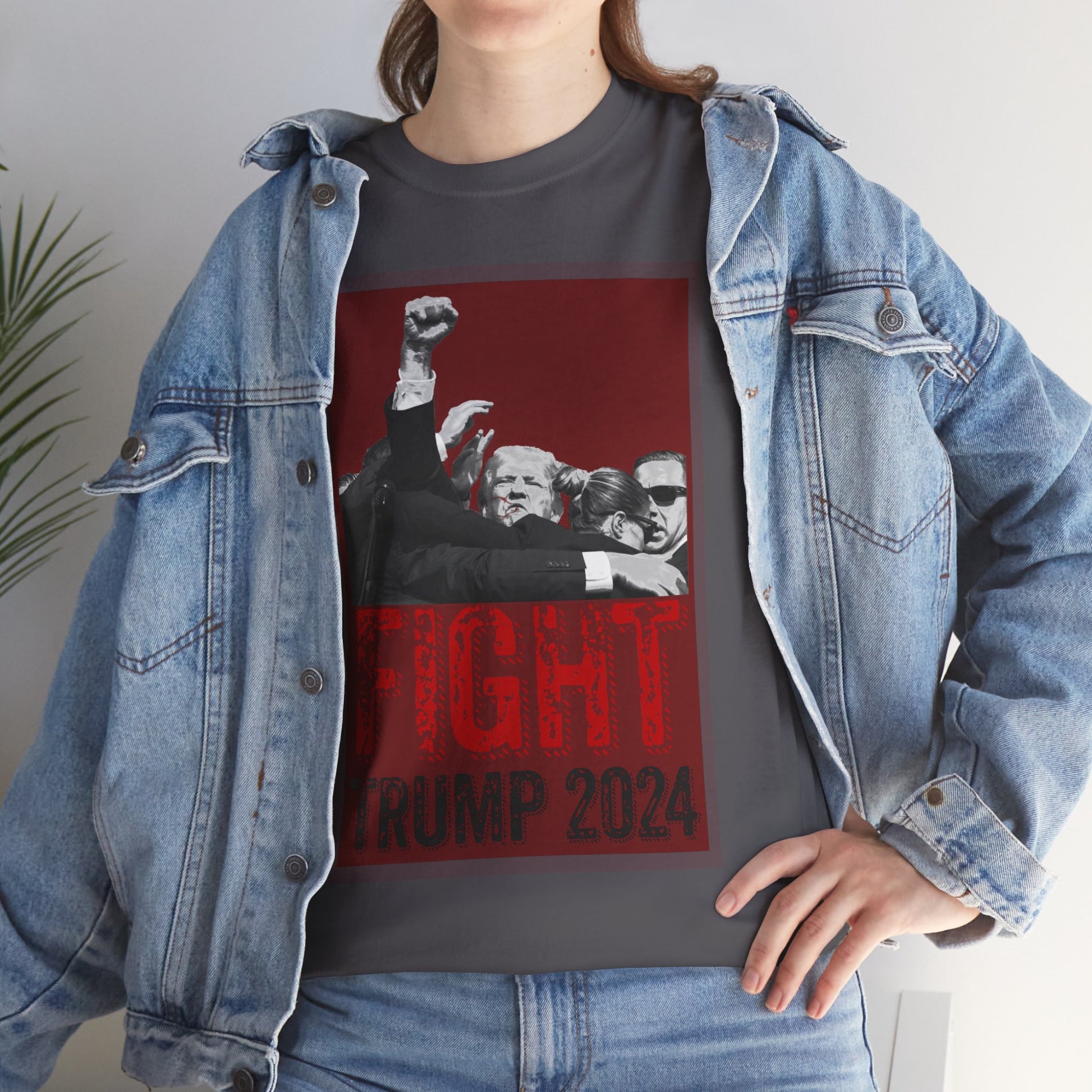 This image shows a person wearing our charcoal colored Fight! Trump 2024 t-shirt with a graphic design of former President Donald Trump raising his fist, surrounded by his secret service, set against a red background. The text “FIGHT” in distressed red font and “TRUMP 2024” in black is printed below the image. The person is also wearing a light blue denim jacket and blue jeans, with one hand on their hip.