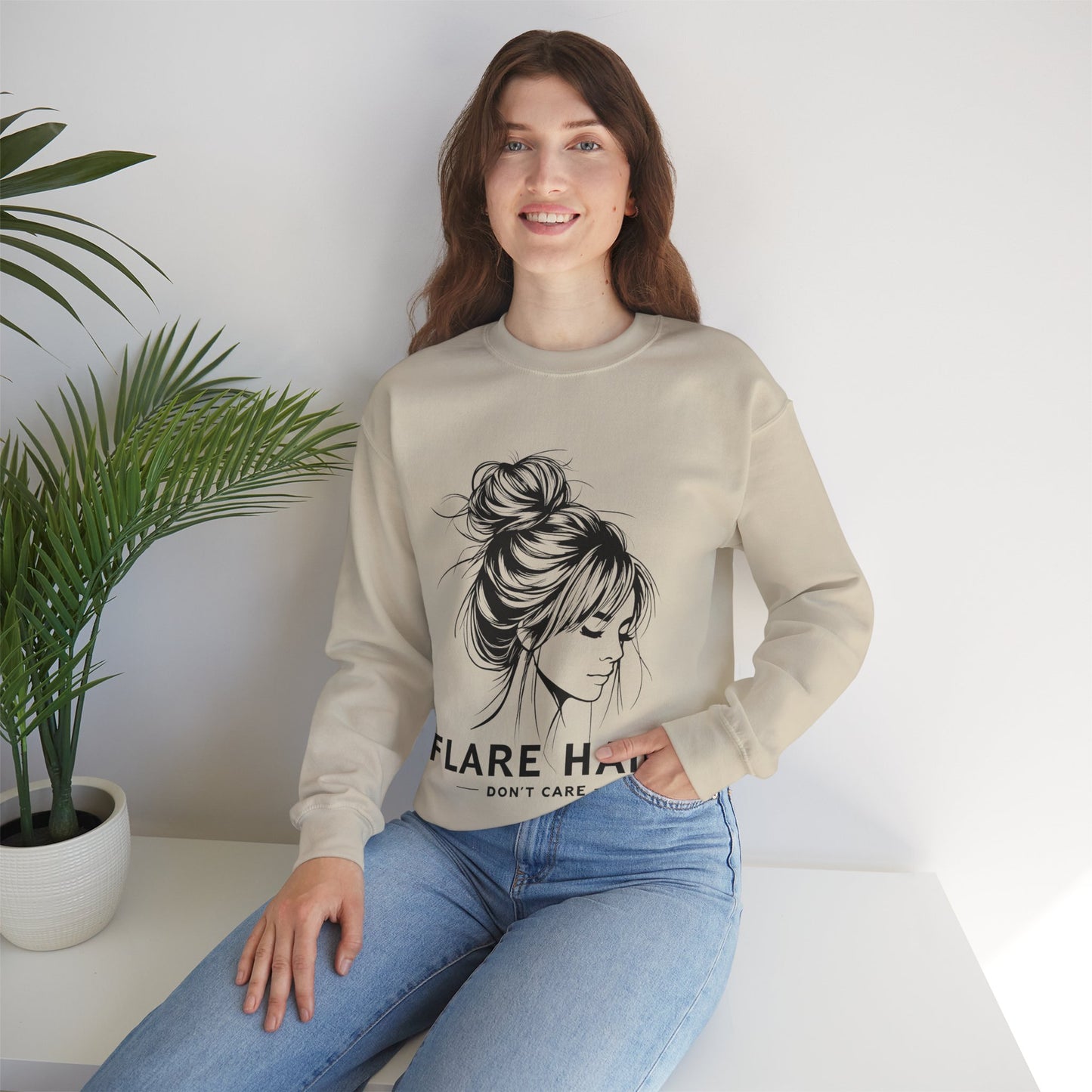 This image features a woman wearing a sand colored crewneck sweatshirt with the text “FLARE HAIR DON’T CARE” printed on the front. The design includes an illustration of a woman with her hair in a messy bun. She is sitting on a white surface with her hand resting on her knee, and there is a potted plant to her left. The background is plain, highlighting the sweatshirt’s design and the casual outfit.