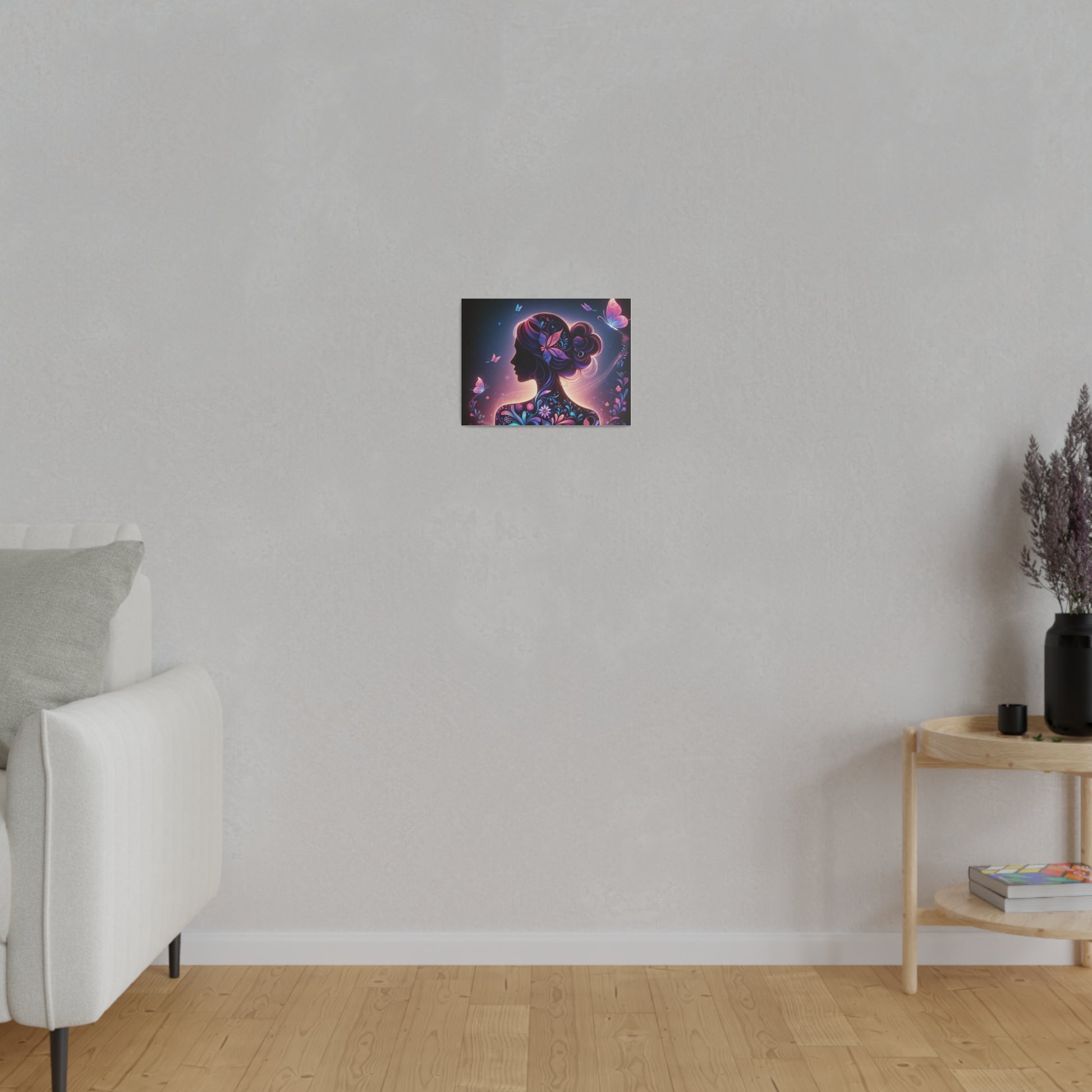 The image shows a room in a minimalist style. On the wall, a canvas artwork titled “Butterfly Bliss. The artwork features the silhouette of a woman in profile with her hair in a bun adorned with vibrant flowers and butterflies in shades of purple, pink, and blue. Butterflies are depicted flying around her. The canvas adds a pop of color to the room, which also includes a white sofa and pillow on the left and a small wooden side table with a black vase with dried lavender and a few books on the right.