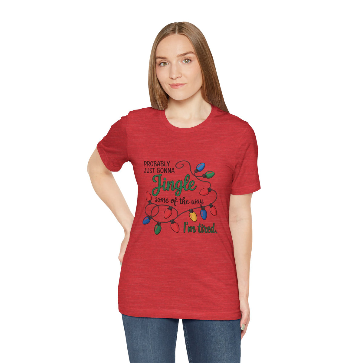 A woman modeling a heather red short-sleeve t-shirt with a Christmas graphic that reads ‘PROBABLY JUST GONNA Jingle some of the way I'm tired.’ with colorful holiday lights, paired with blue jeans against a plain white background.