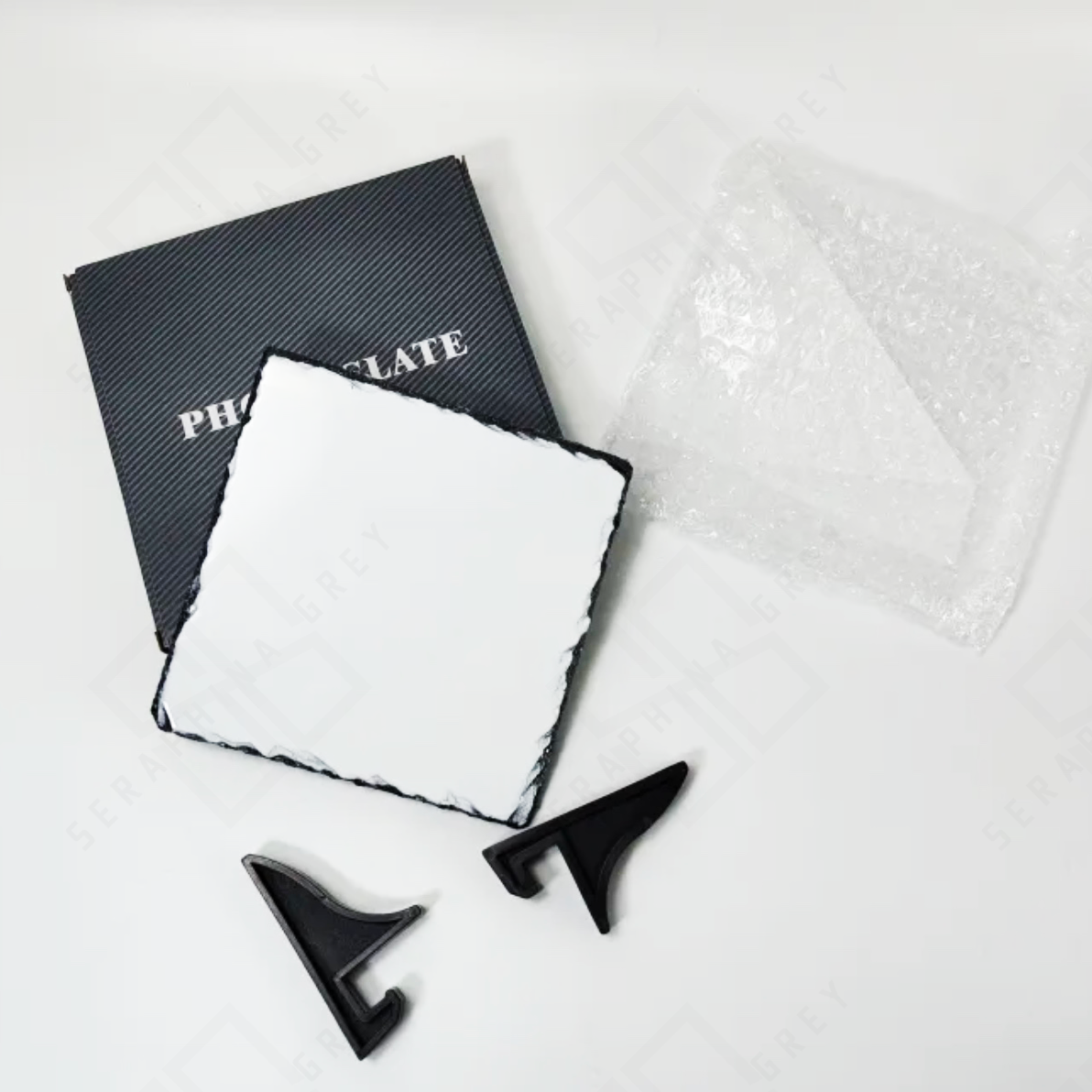 This image shows the components of a custom photo slate set. The set includes a blank white photo slate with a natural, uneven edge, two black stands, a bubble wrap sheet, and a black box labeled “PHOTO SLATE.” The items are arranged on a white surface, highlighting the slate’s rustic edge and the protective packaging materials included in the set. The watermark with “Seraphina Grey” is faintly visible across the background.