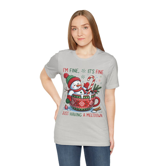 A woman modeling a silver short-sleeve t-shirt with a Christmas graphic of a snowman melting into a mug and the text ‘I'M FINE, IT'S FINE JUST HAVING A MELTDOWN’, paired with blue jeans against a plain white background.