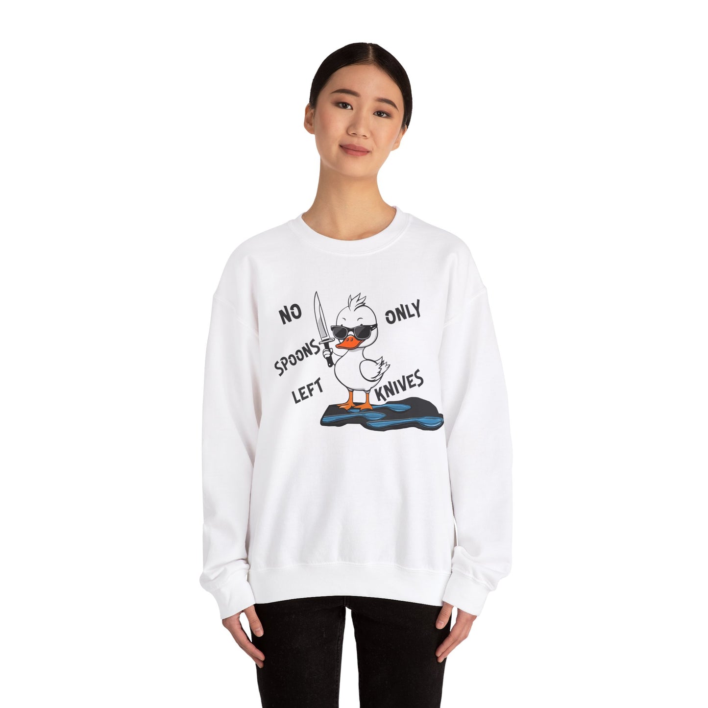 This image shows a woman wearing a white crewneck sweatshirt with a cartoon design of a duck holding a knife, standing in a puddle of spoons. The text around the duck reads “NO SPOONS LEFT, ONLY KNIVES.” The woman is standing against a plain white background, allowing the sweatshirt’s design to be clearly visible. She is wearing black pants and has a slight smile on her face.