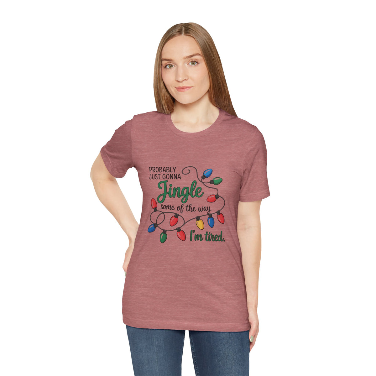 A woman modeling a heather mauve short-sleeve t-shirt with a Christmas graphic that reads ‘PROBABLY JUST GONNA Jingle some of the way I'm tired.’ with colorful holiday lights, paired with blue jeans against a plain white background.