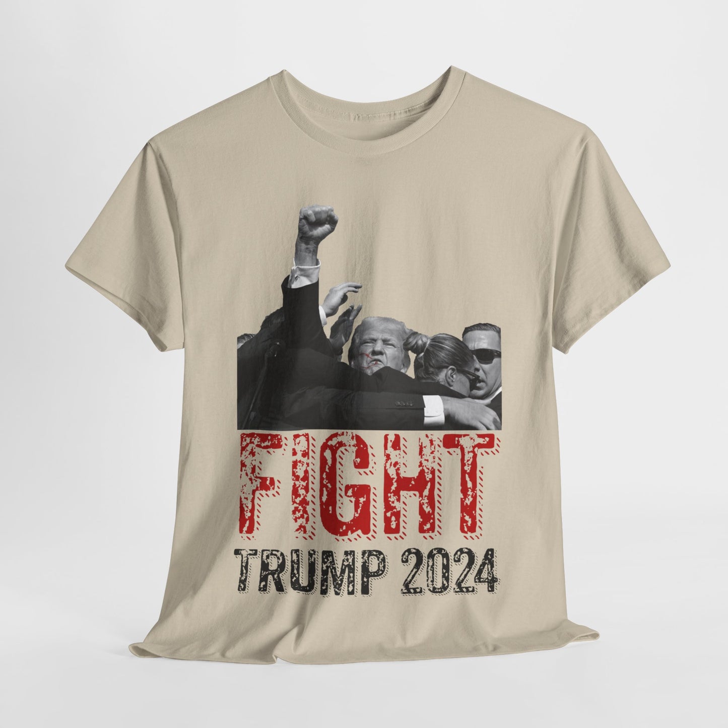 This image displays a sand-colored, unisex, heavy cotton t-shirt with a graphic design on the front. The design features a black-and-white photograph of former President Donald Trump raising his fist, being shielded by brave men and women. Below the photograph, the words “FIGHT” and “TRUMP 2024” are printed in bold letters, with “FIGHT” in a distressed red font and “TRUMP 2024” in black. The t-shirt is laid out on a plain white background, showcasing the design prominently.