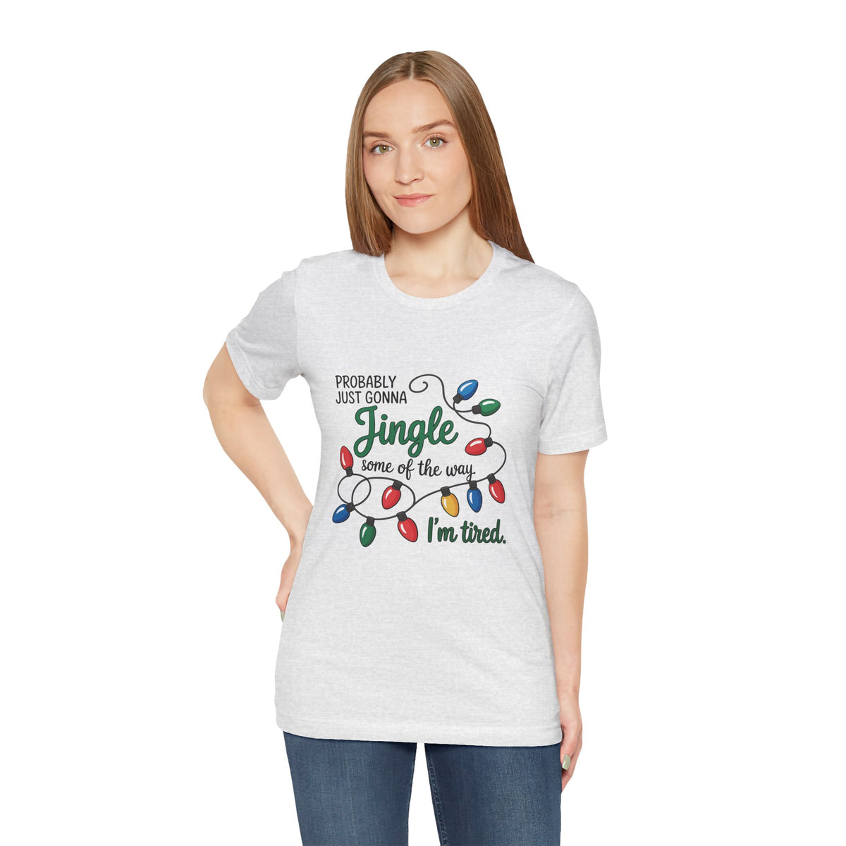 A woman modeling an ash short-sleeve t-shirt with a Christmas graphic that reads ‘PROBABLY JUST GONNA Jingle some of the way I'm tired.’ with colorful holiday lights, paired with blue jeans against a plain white background.