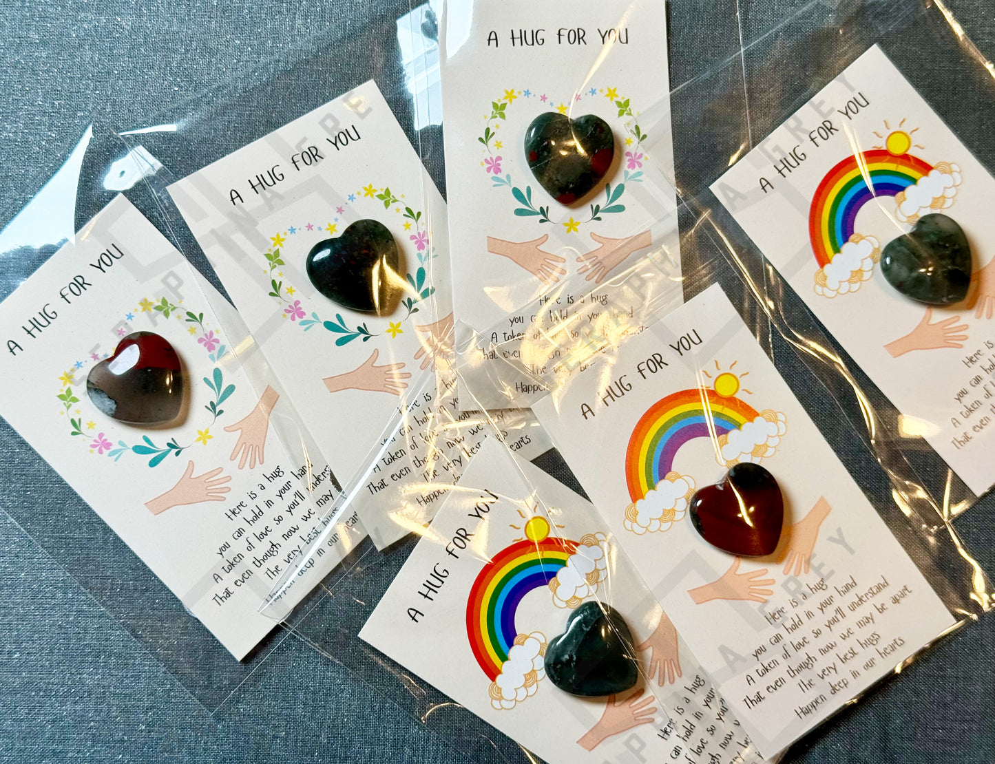 A collection of cards, each featuring an African bloodstone heart and the text ‘A Hug For You.’ The cards include different designs, such as a floral wreath and a rainbow with clouds and sun. Each card has an illustration of two hands reaching out, with a message expressing the warmth and comfort of a hug, even when apart. The cards are individually packaged in plastic sleeves.