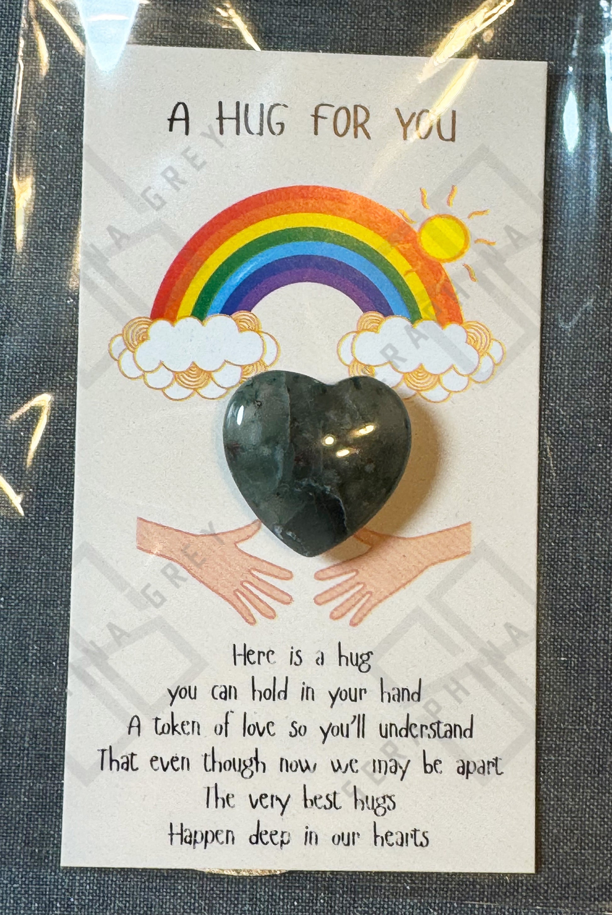 A card featuring an African bloodstone heart with the text ‘A Hug For You’ at the top. The design includes a colorful rainbow with clouds and a shining sun above the heart, along with an illustration of two hands reaching out. The message below the heart expresses that the heart stone is a token of love, providing comfort even when apart. The card is wrapped in a plastic sleeve.