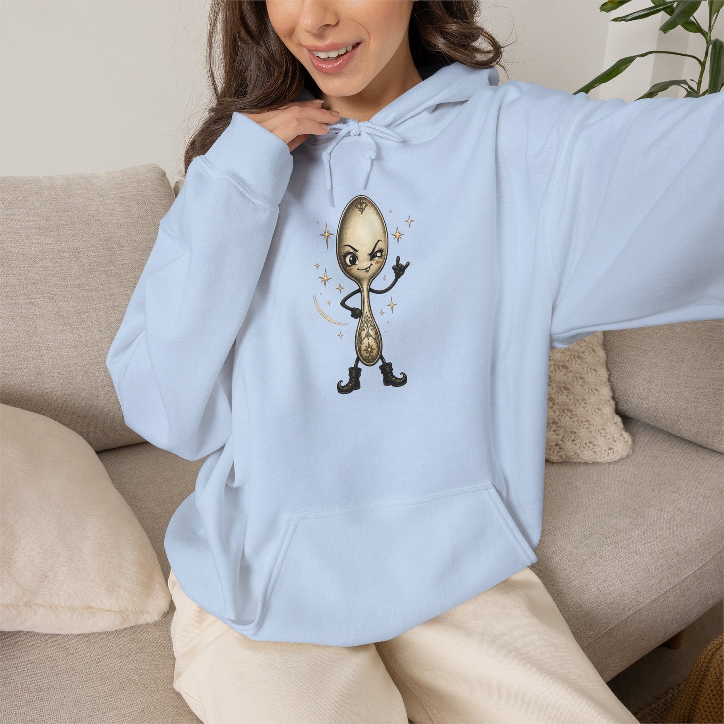 Light blue pullover hoodie worn by a model, featuring a whimsical vintage-style cartoon spoon making a rock hand gesture with small star accents, shown in a casual indoor setting.