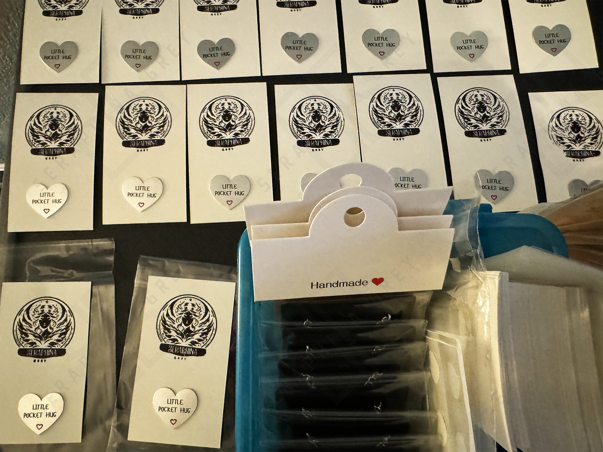 An assortment of ‘Little Pocket Hug’ cards in the making, arranged neatly on a table. Each card features a small heart with the text ‘Little Pocket Hug’ and a small red heart symbol below. Some cards display a black and white intricate design at the top, while others are plain. A container labeled ‘Handmade’ is seen holding multiple packaged cards, indicating preparation for distribution