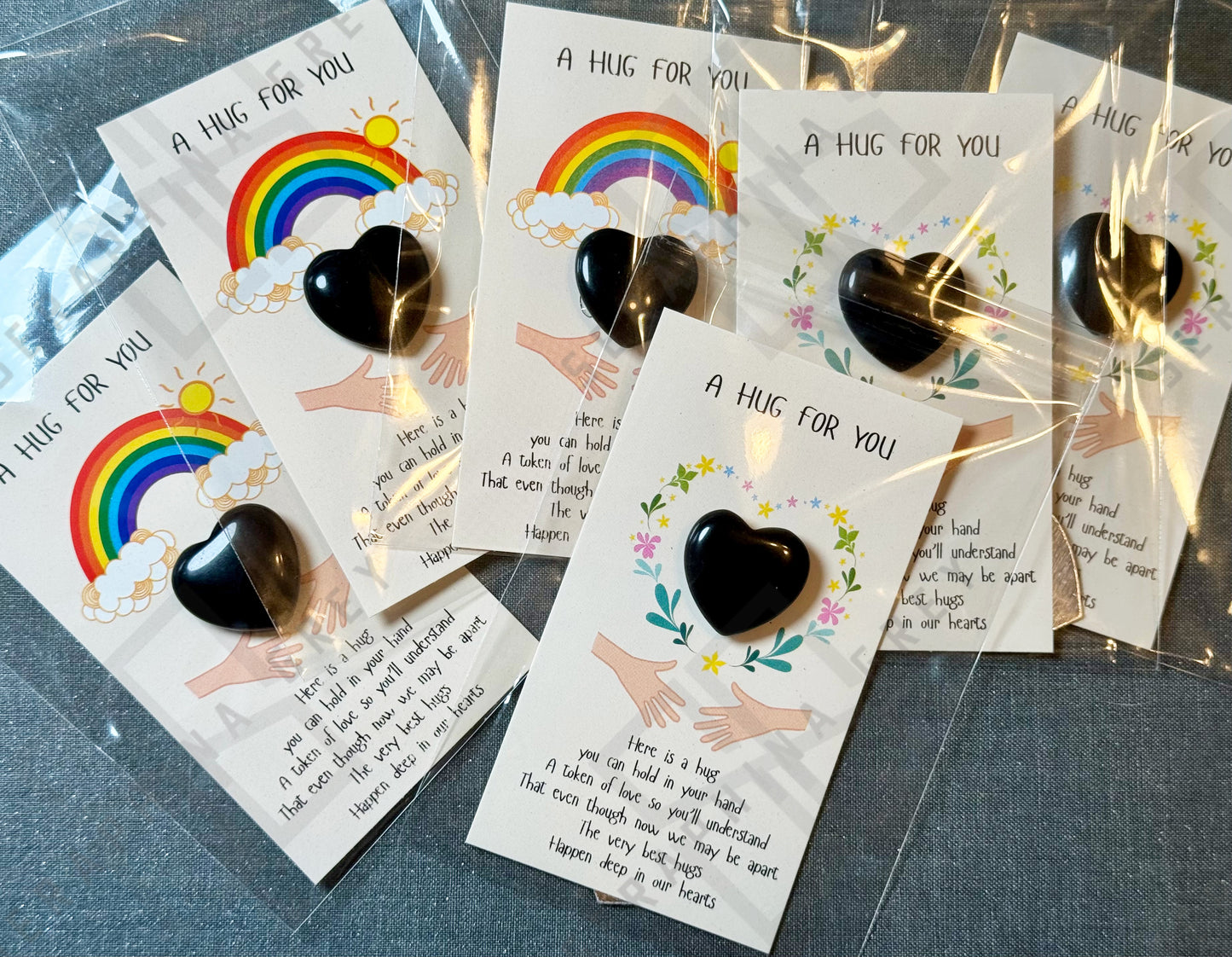 A collection of cards, each featuring a black obsidian heart and the text ‘A Hug For You.’ The cards have various designs, including a rainbow with clouds and sun, and a floral wreath. Each card has an illustration of two hands reaching out and a message expressing the warmth and love of a hug, even when apart. The cards are individually wrapped.