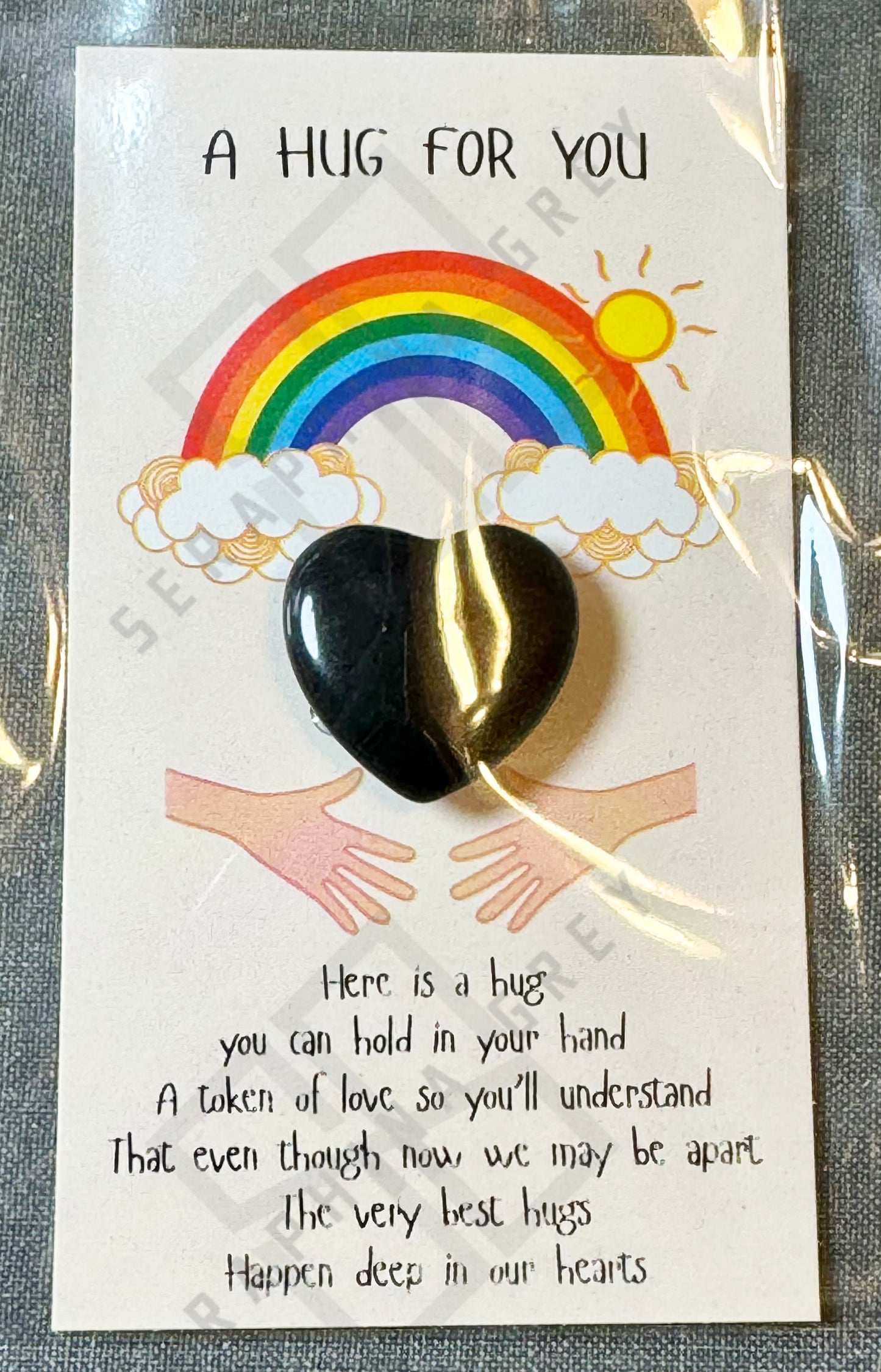 A card featuring a black obsidian heart with the text ‘A Hug For You’ at the top. The design includes a colorful rainbow with clouds and a shining sun above the heart, and an illustration of two hands reaching out. Below the heart, there is a heartfelt message expressing that a hug can be held in your hand as a token of love, even when apart. The card is wrapped in a plastic sleeve.