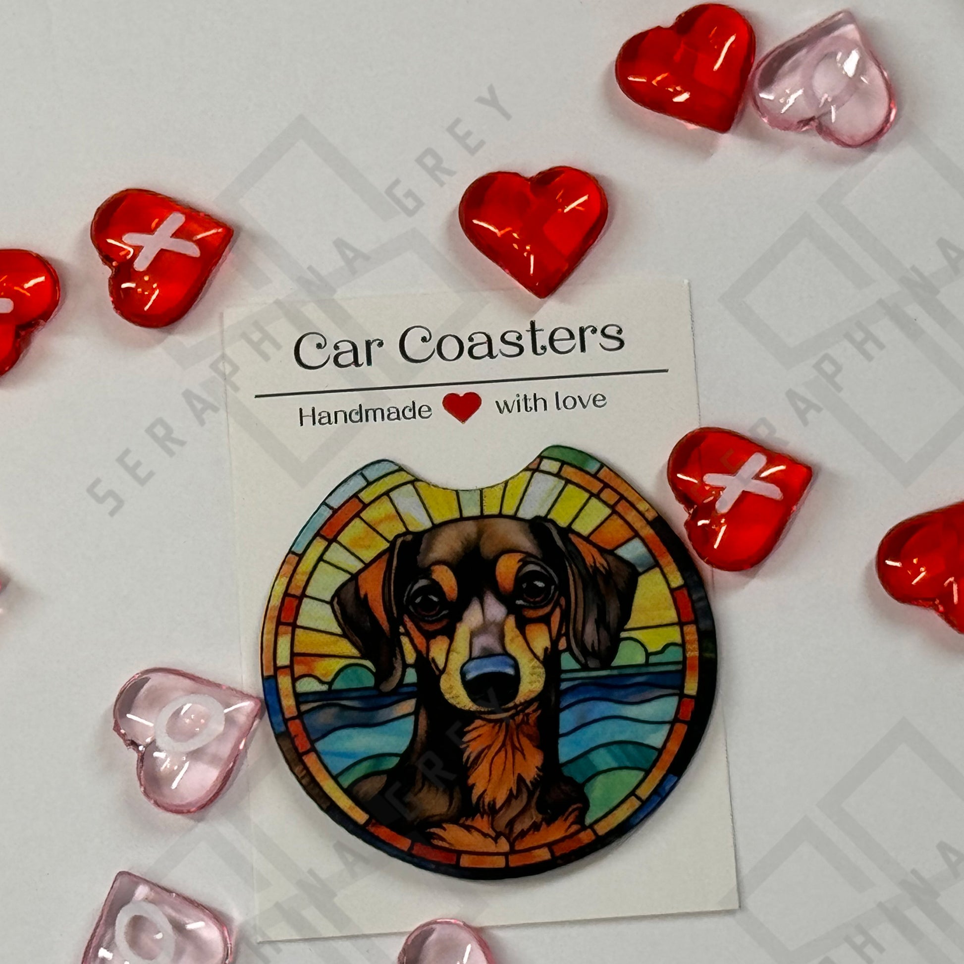 A single car coaster featuring an artistic design of a black and tan dachshund dog against a vibrant, stained-glass-style background. The coaster is presented with a label that reads ‘Car Coasters - Handmade with love.’ The display is surrounded by red and pink heart-shaped decorations.