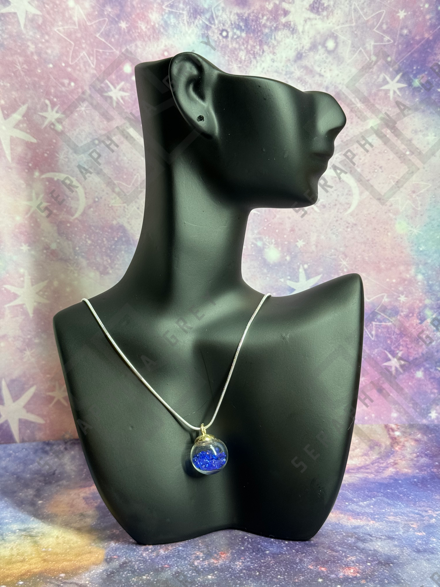 This image features a glass ball pendant necklace displayed on a black mannequin bust. The pendant is a glass ball with a gold-colored cap, filled with blue crystals, hanging from a thin silver chain. The background is a colorful, galaxy-themed pattern with stars and nebula-like designs, enhancing the visual appeal of the necklace.